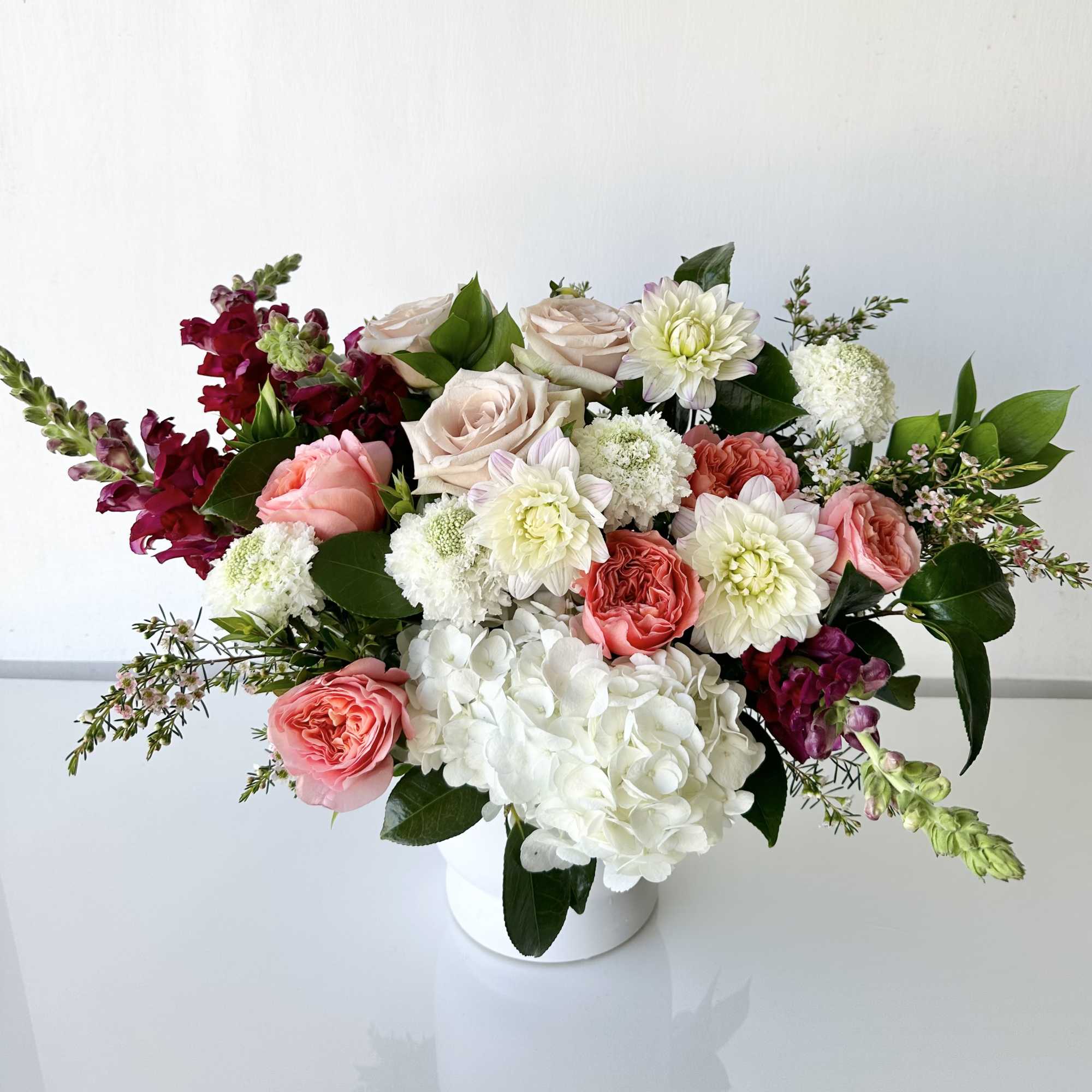 An arrangement of fresh-cut flowers includes roses, dahlia, scabiosa, hydrangea, and snapdragon