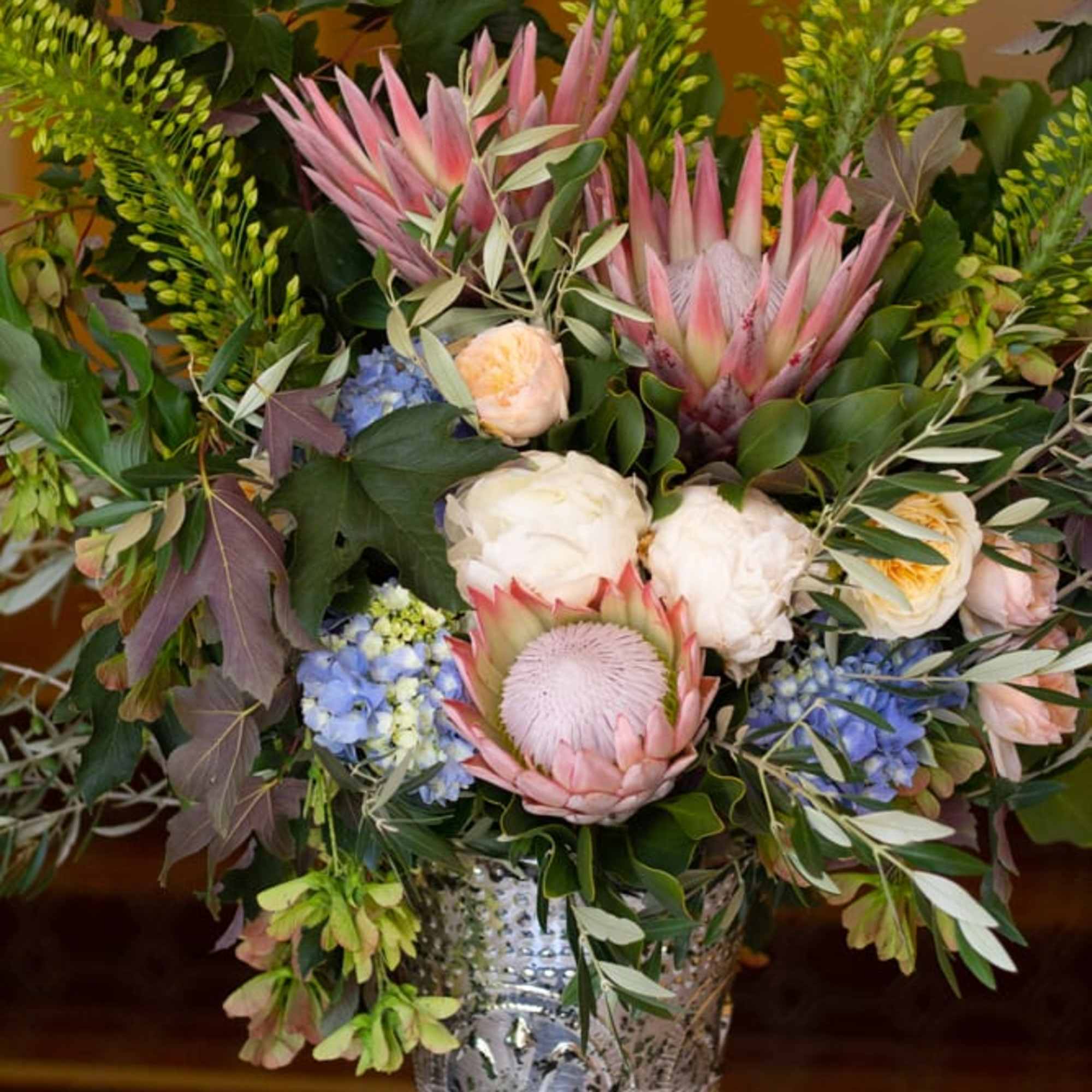 Large mixed floral arrangement in a silver vase with pink protea and blue blooms