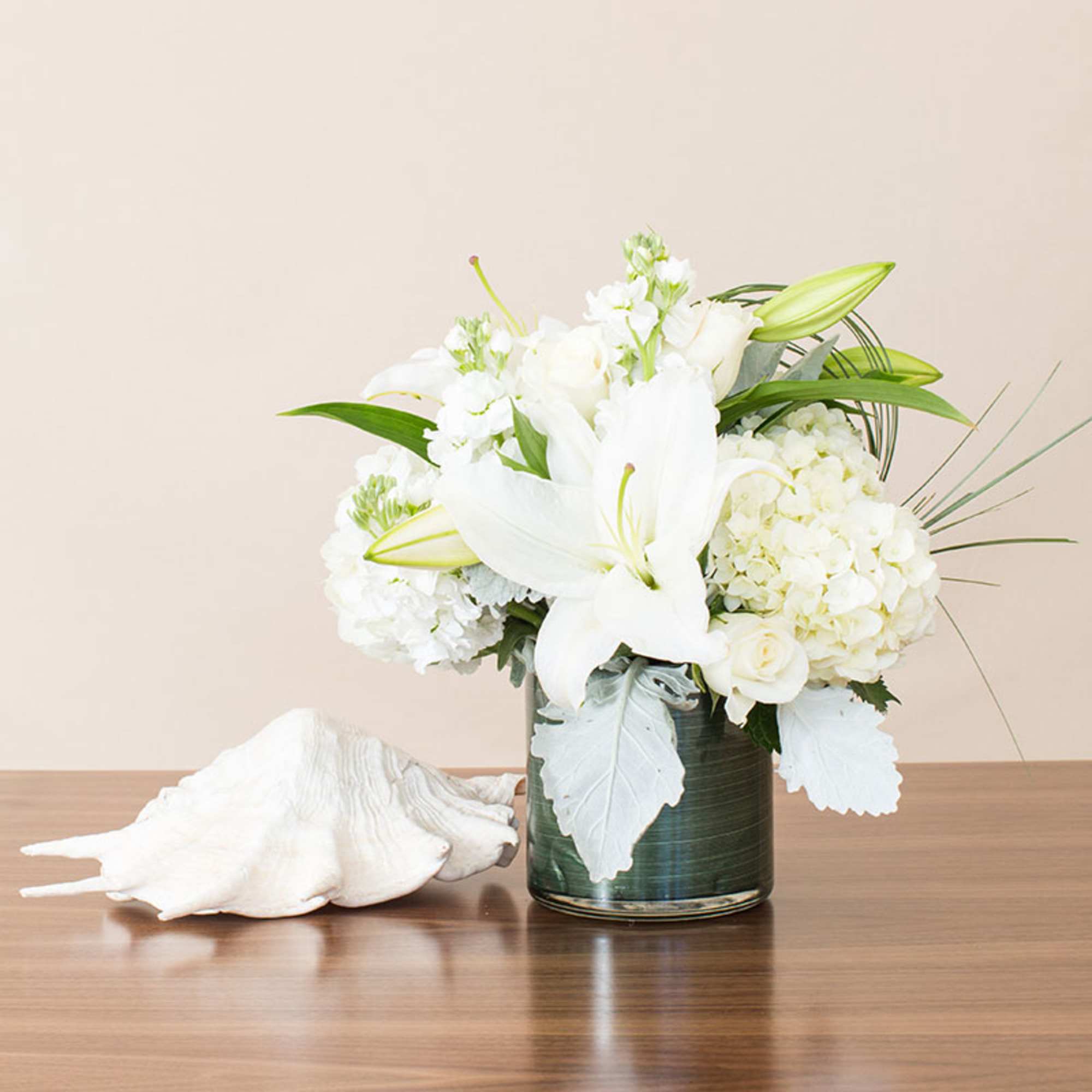 White lilies, roses, and hydrangeas in a glass vase beside a large white seashell.