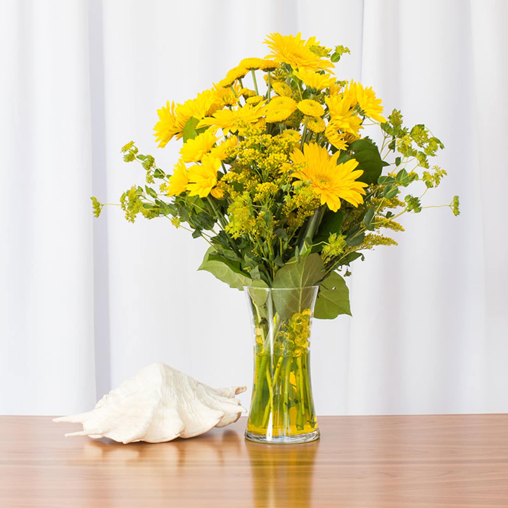 Tall clear vase of bright yellow daisies and gerbera daisies on a table with a white seashell beside it