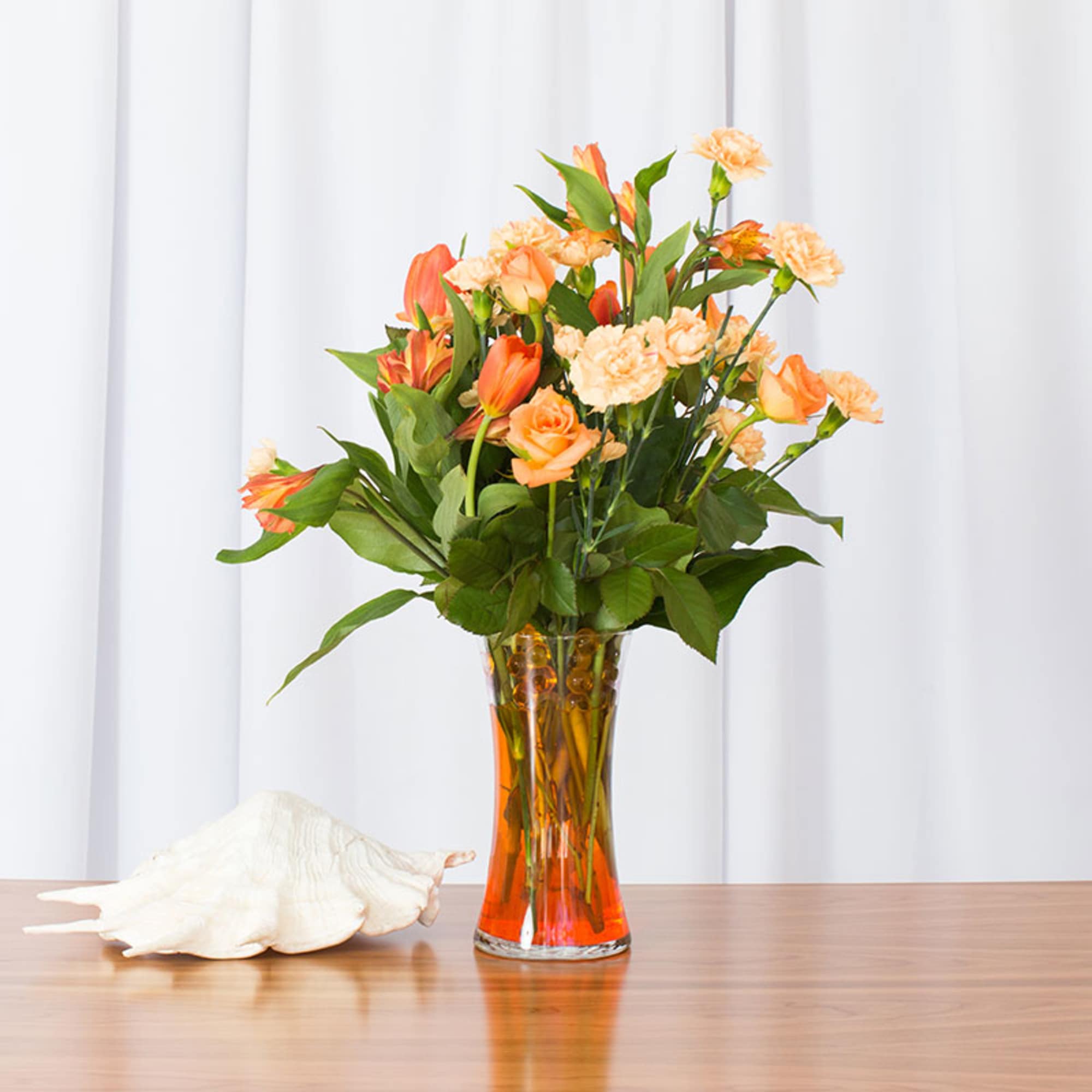 Tall arrangement of orange roses, tulips, and peach carnations in a clear vase on a table