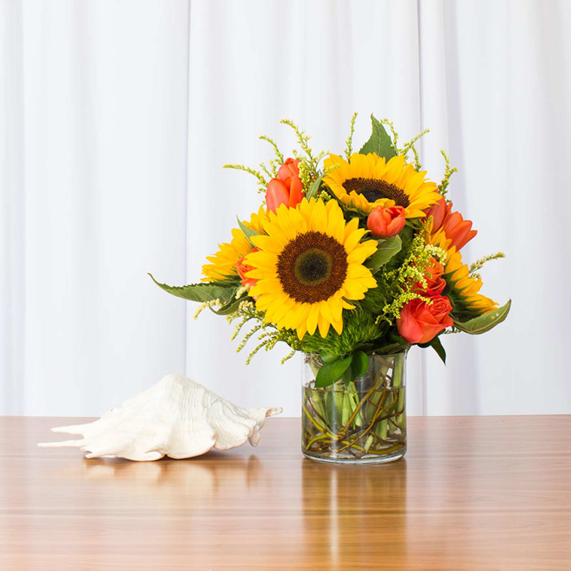 Bright bouquet of yellow sunflowers and orange roses in a clear glass vase beside a white seashell.