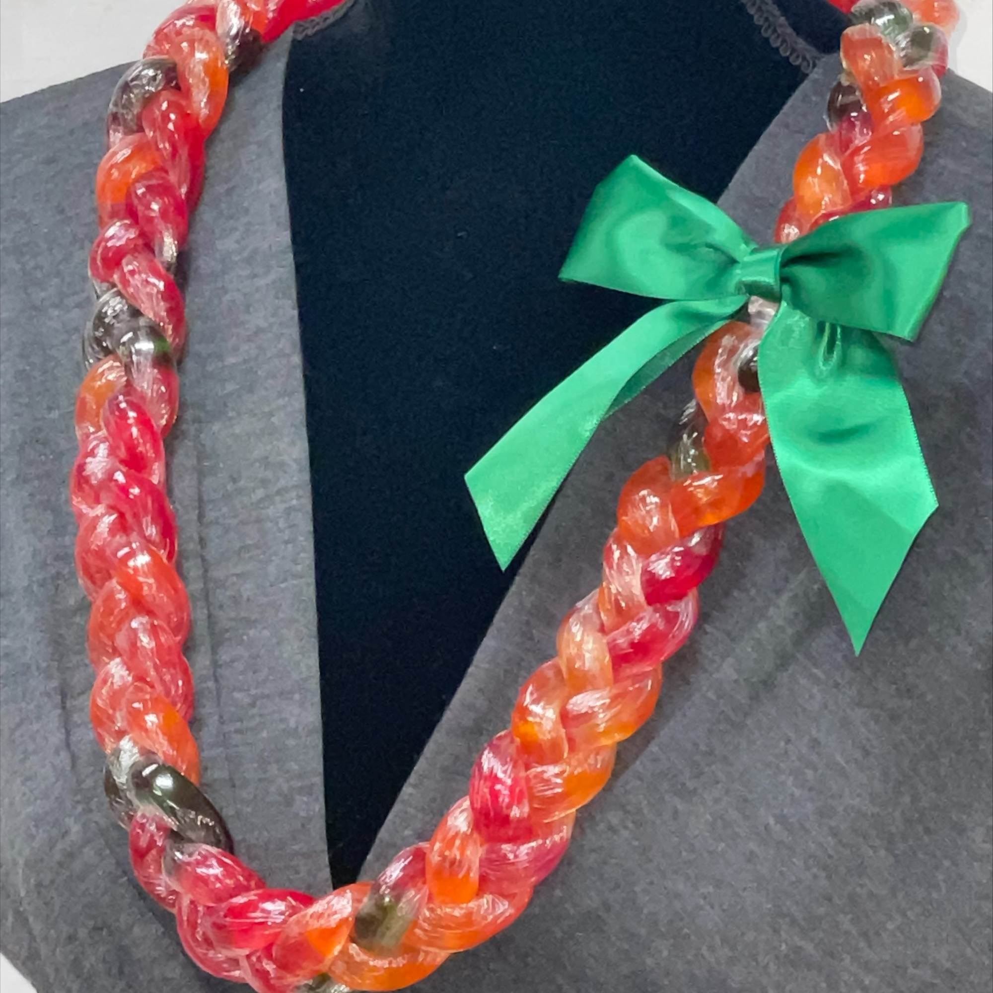 Braided candy lei in red and orange hues with a green satin bow on a display bust.