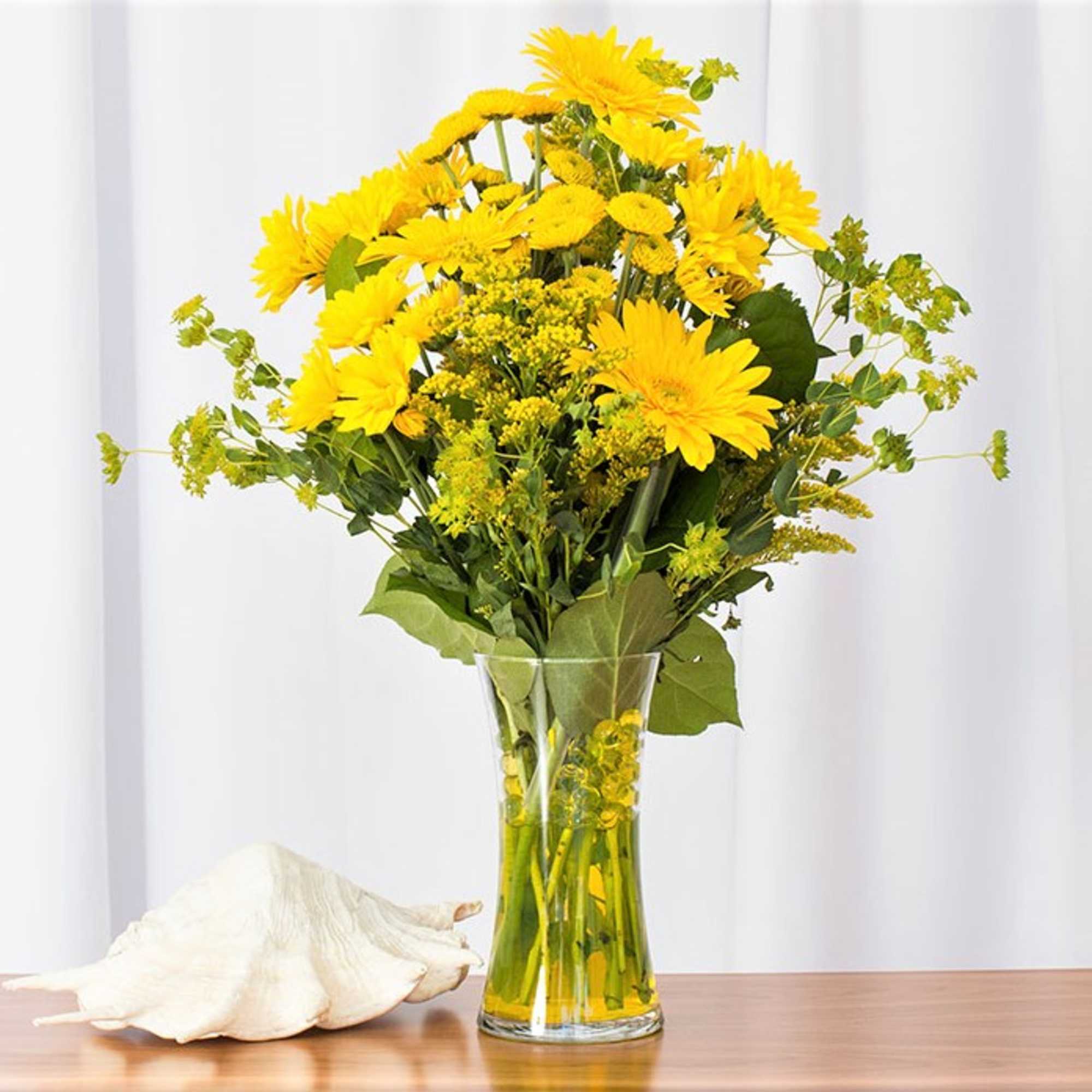 Tall bouquet of yellow gerbera daisies in a clear glass vase beside a white seashell.
