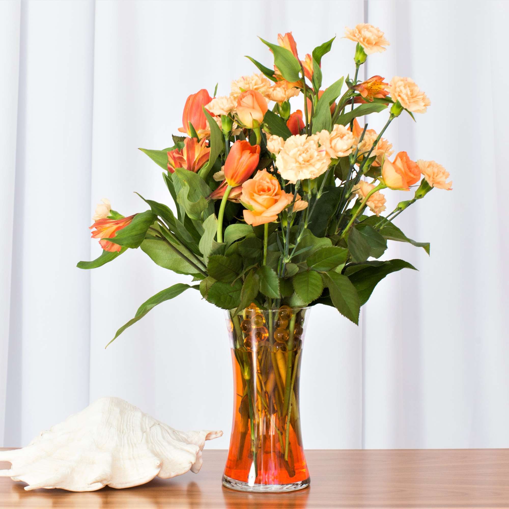 Tall arrangement of orange and peach roses, carnations, and other blooms in a clear glass vase with orange water.