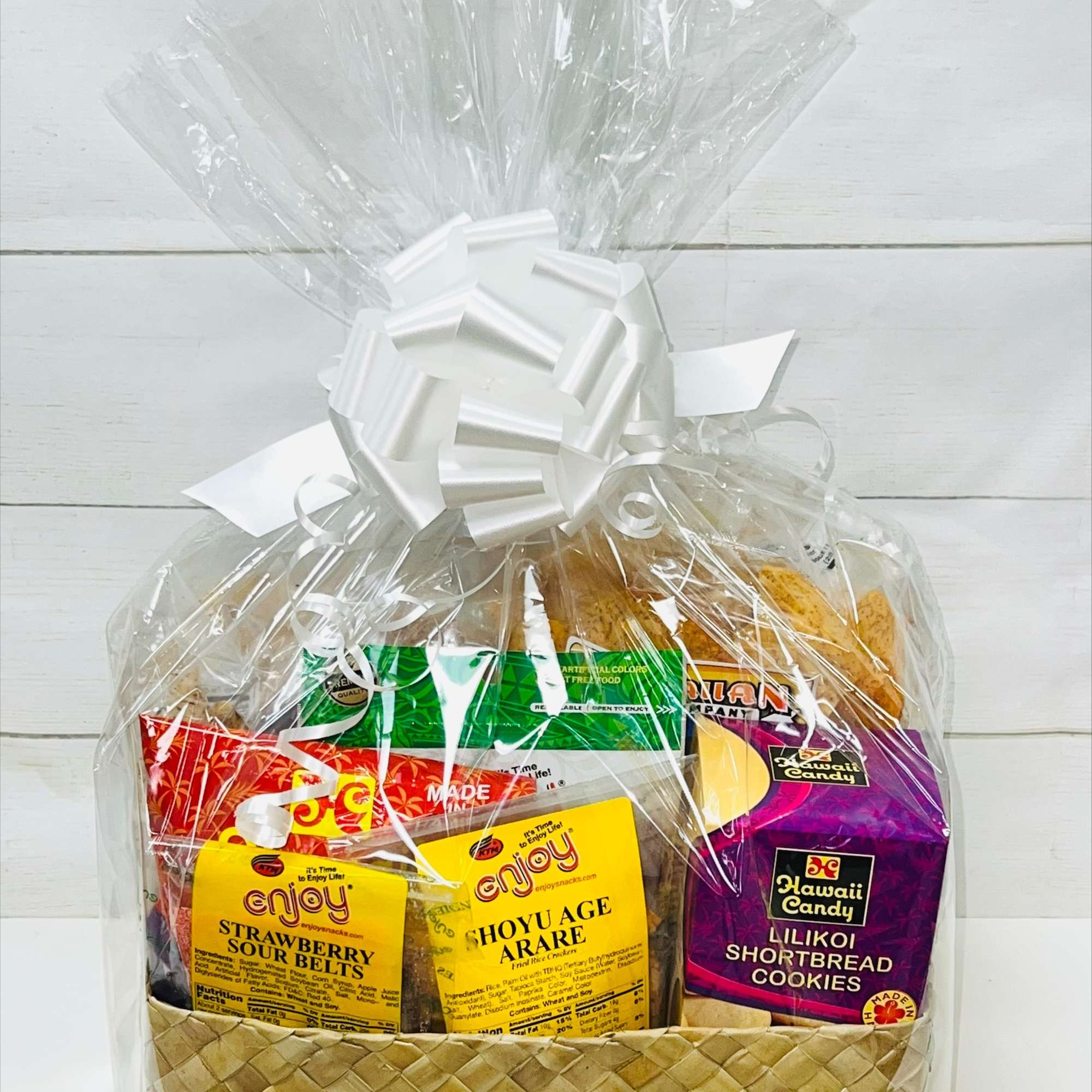 Woven gift basket wrapped in clear cellophane, filled with assorted colorful snack packages and topped with a white bow.