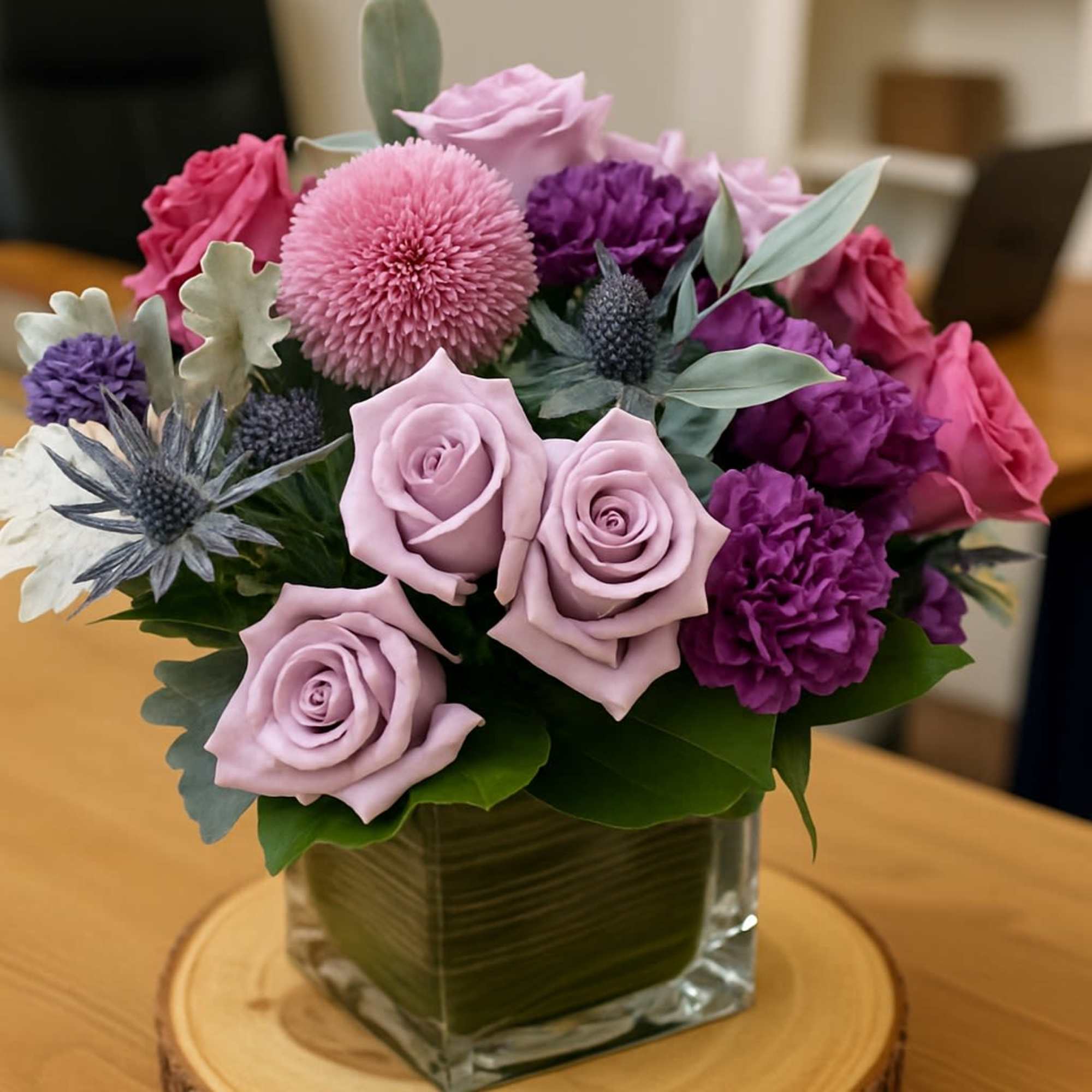 Compact arrangement of pink and purple flowers in a square glass vase on a wood slice.