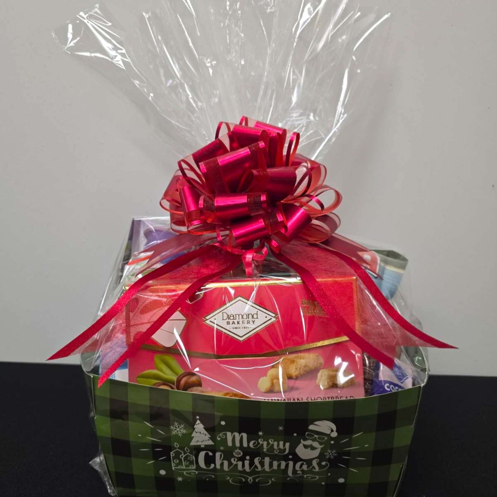 Christmas gift basket wrapped in clear cellophane with red bow and snacks in a green box.