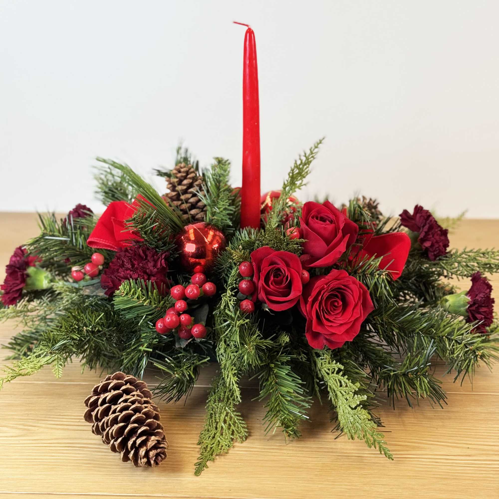 Long centerpiece with red roses, a red taper candle, evergreens, pinecones, and red berries.