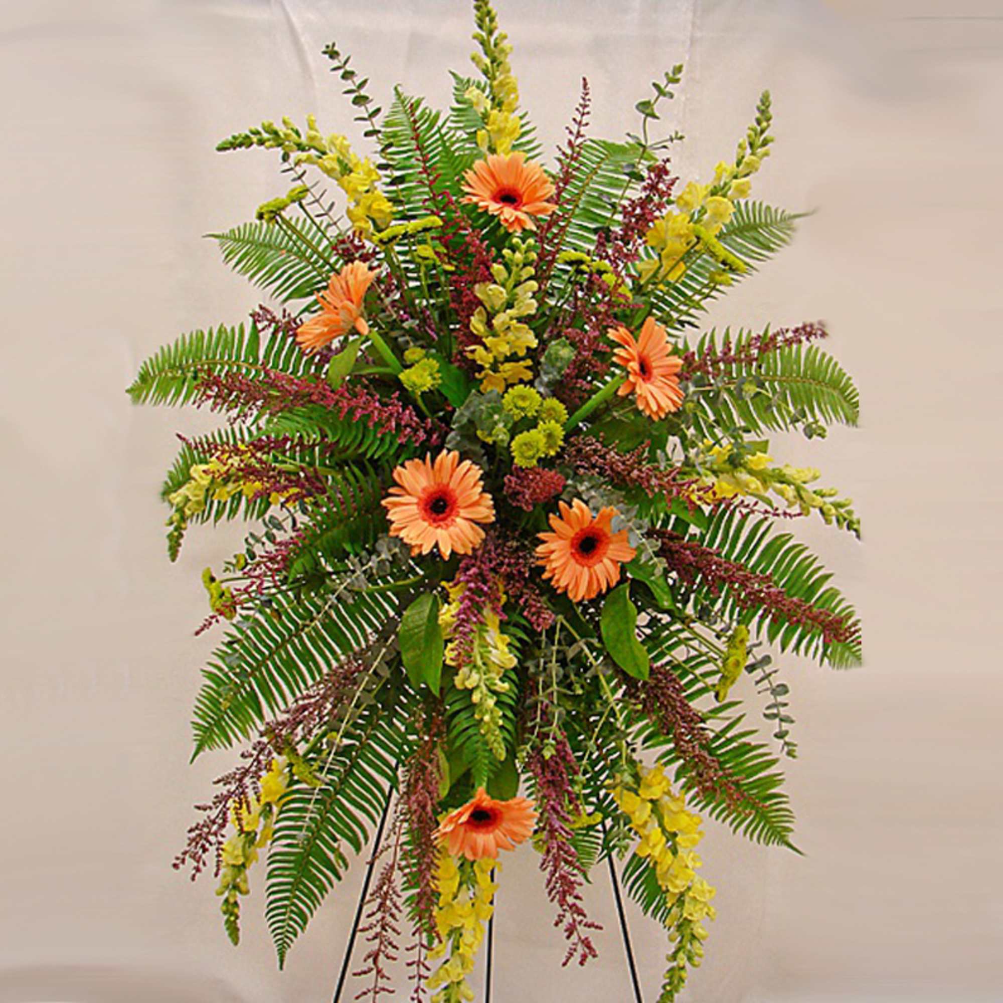 Tall standing spray with orange gerbera daisies, yellow flowers, and ferns on a metal easel.