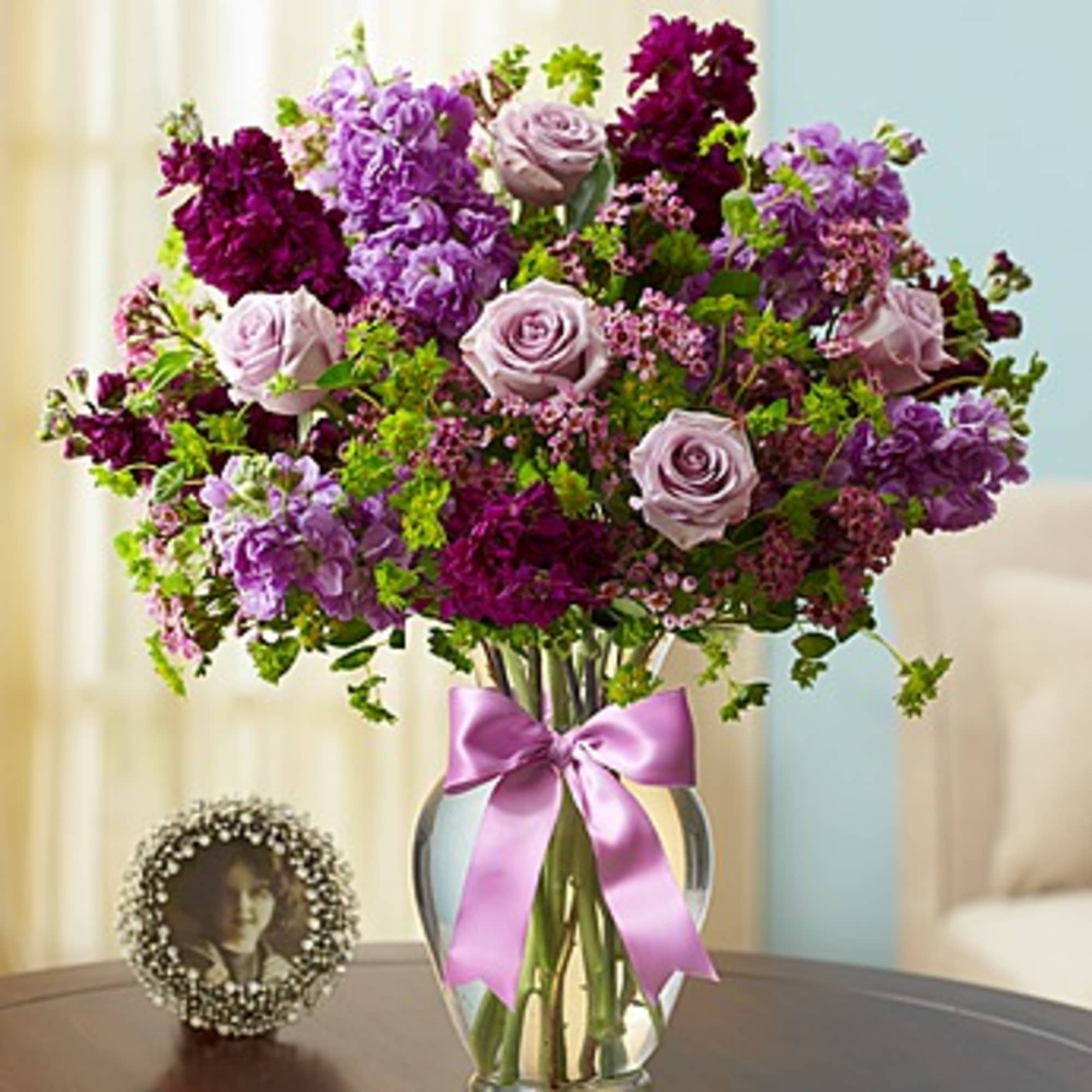 Purple and lavender flower arrangement in a glass vase with a pink ribbon
