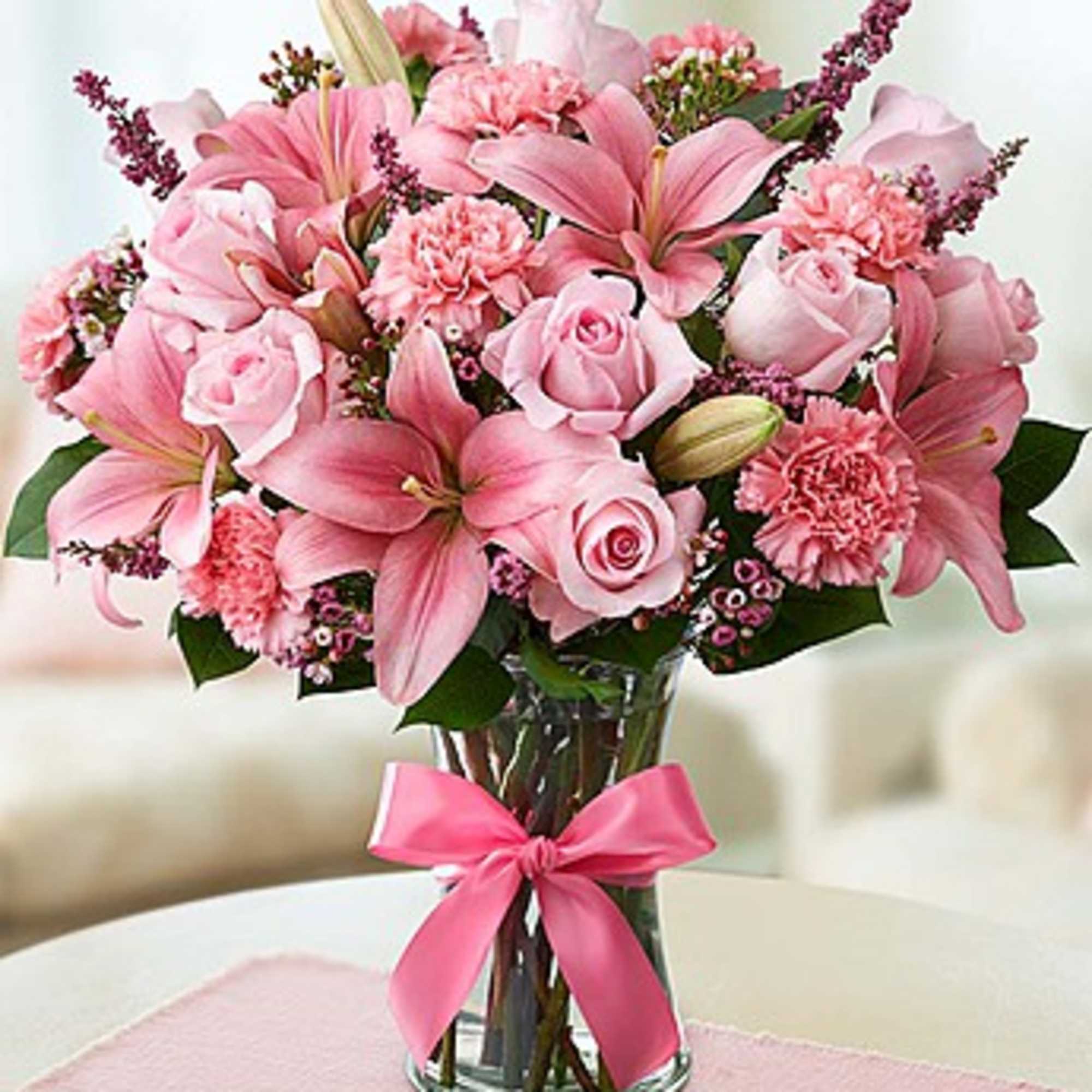 Pink lilies and roses arranged in a glass vase with a pink ribbon.