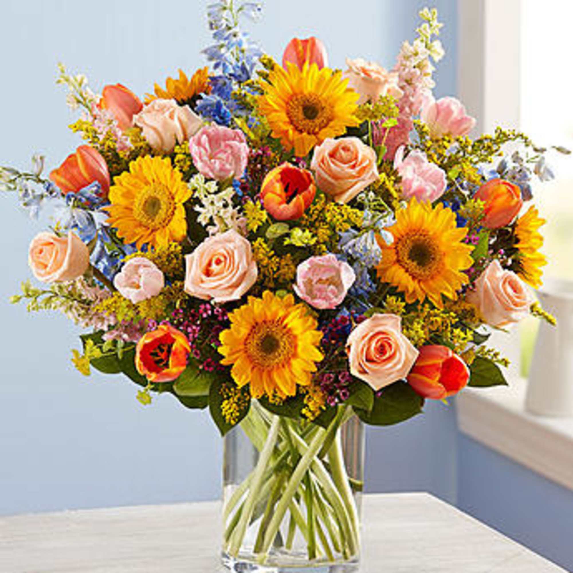 Mixed bouquet of sunflowers, roses, and tulips in a clear glass vase