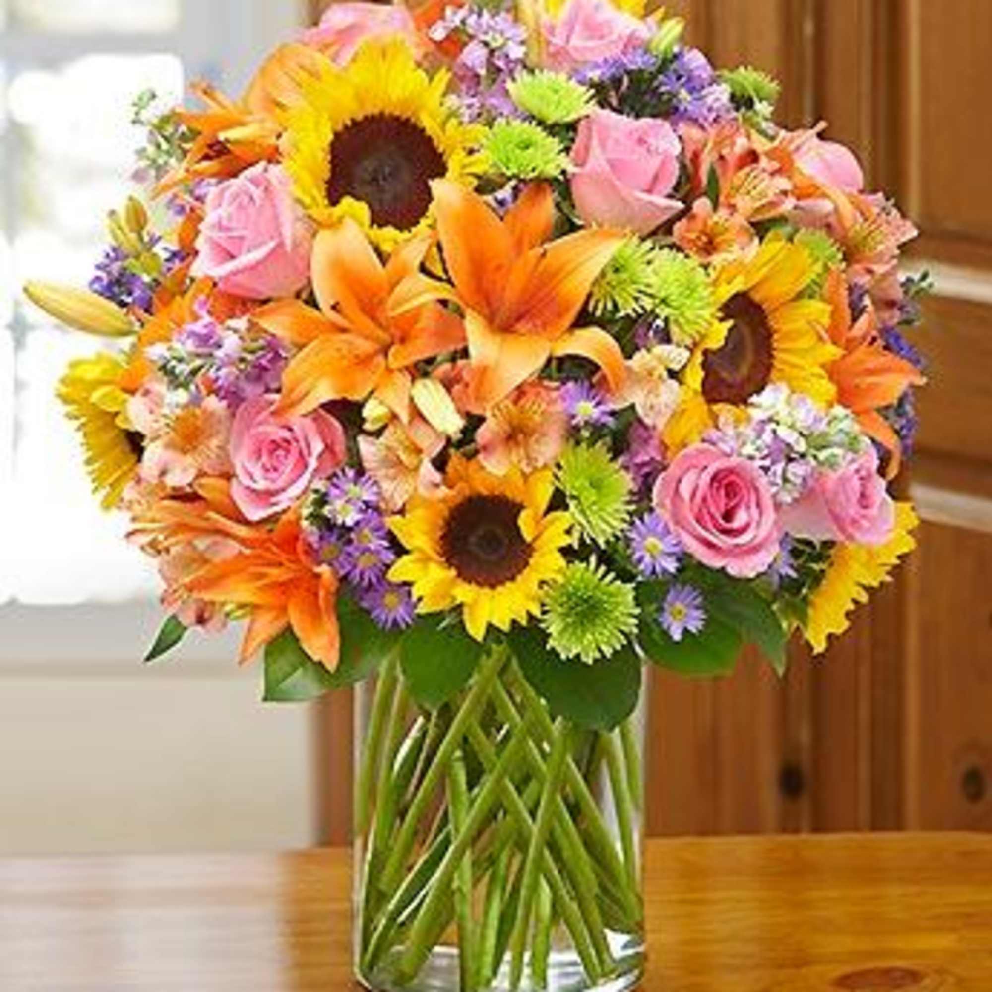 Mixed bouquet of sunflowers, pink roses, lilies, and purple flowers in a glass vase