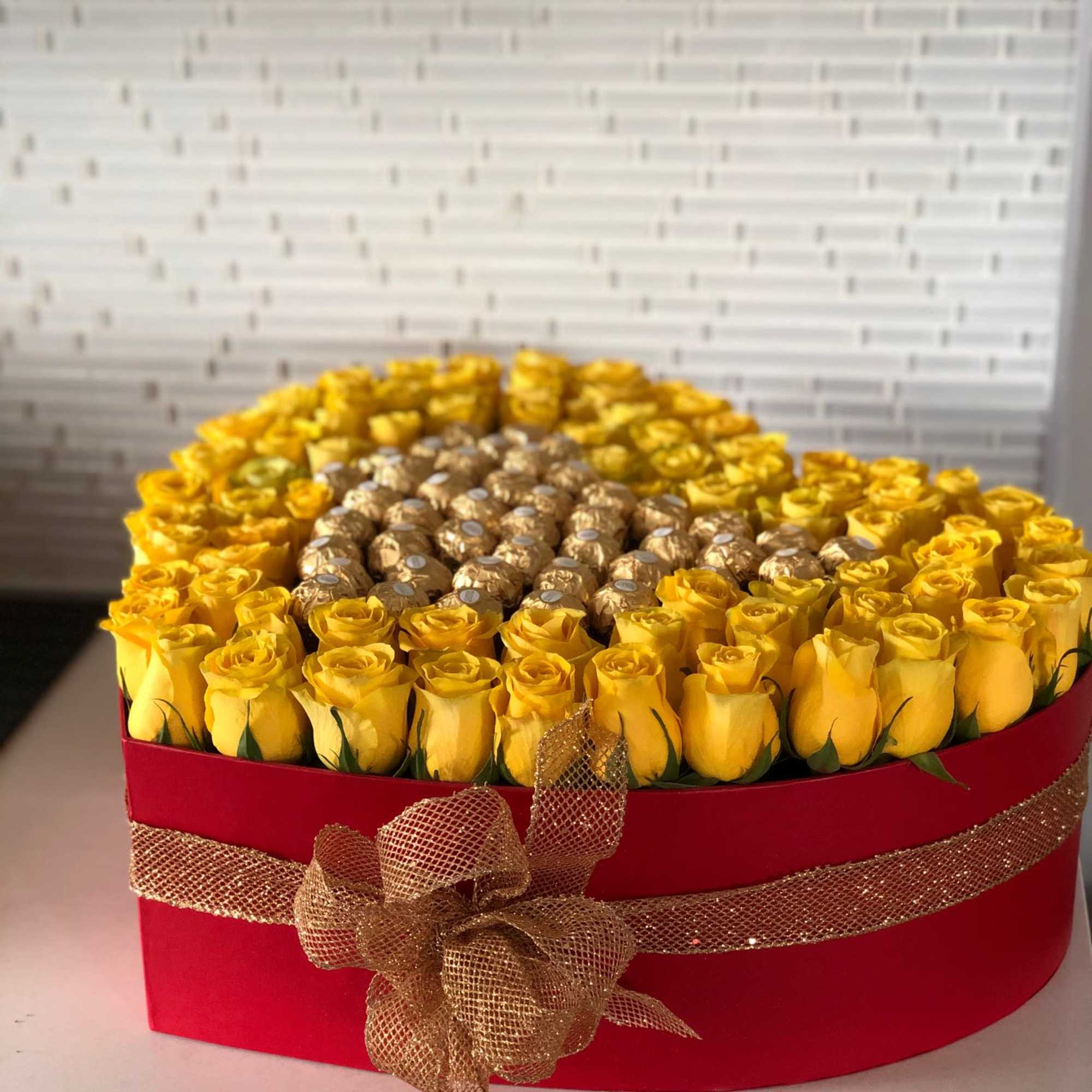 Yellow roses arranged in a red hat box with gold ribbon and chocolates in the center