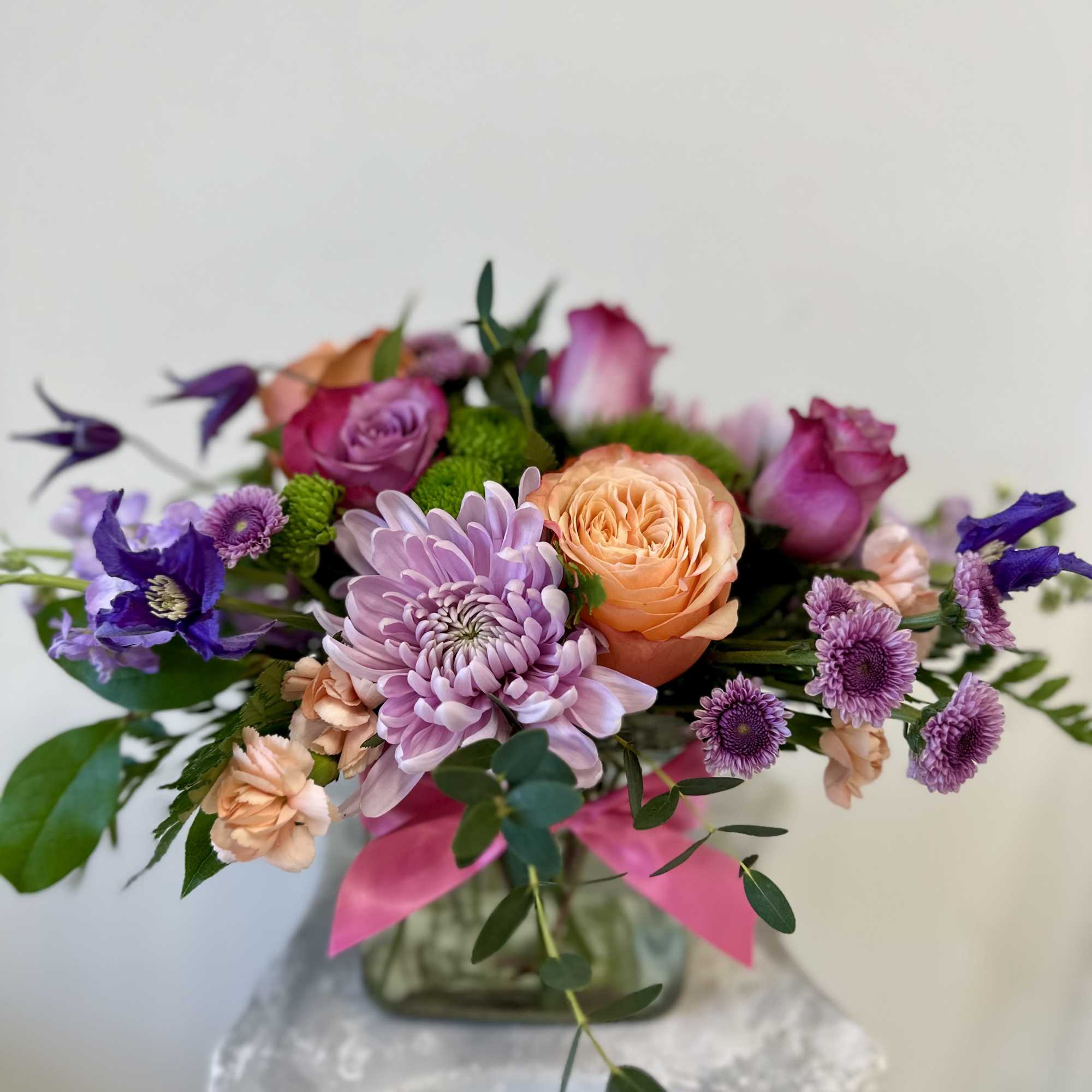 Low mixed arrangement of pink, purple, and peach flowers in a glass cube vase with a pink ribbon.