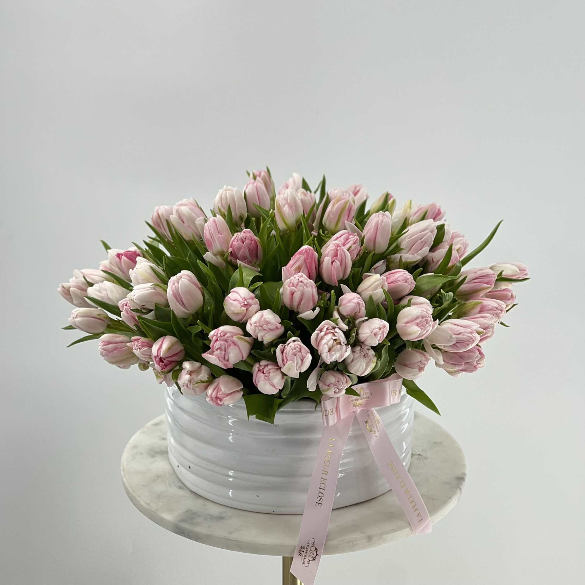Pink tulips arranged in a white round container with a pink ribbon
