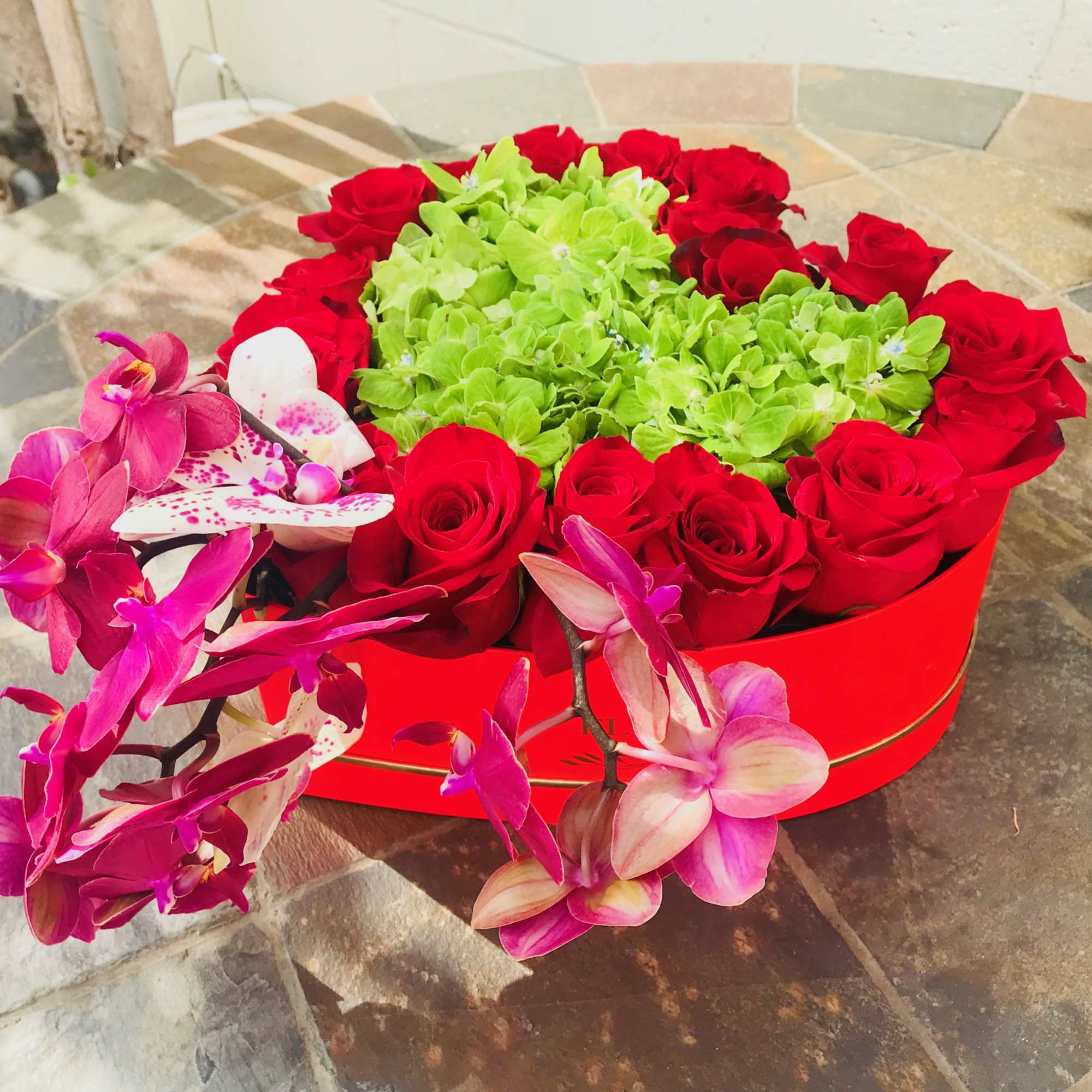 Red roses and green hydrangeas in a red box with pink orchids draped over the side