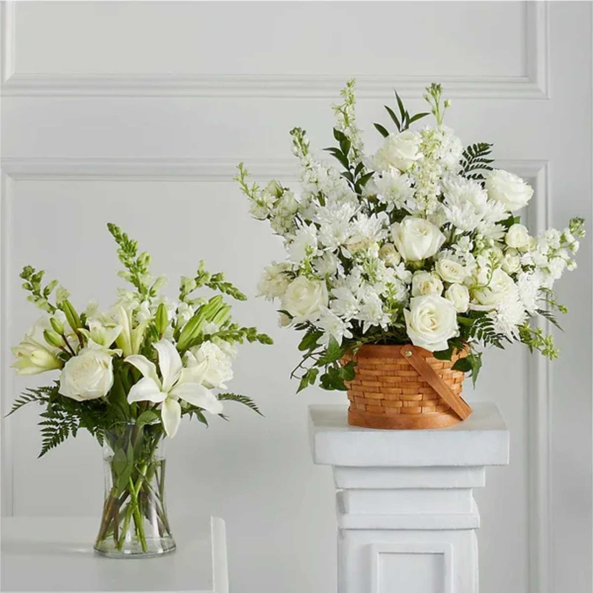Two white floral arrangements with roses and lilies, one in a glass vase and one in a woven basket on a pedestal