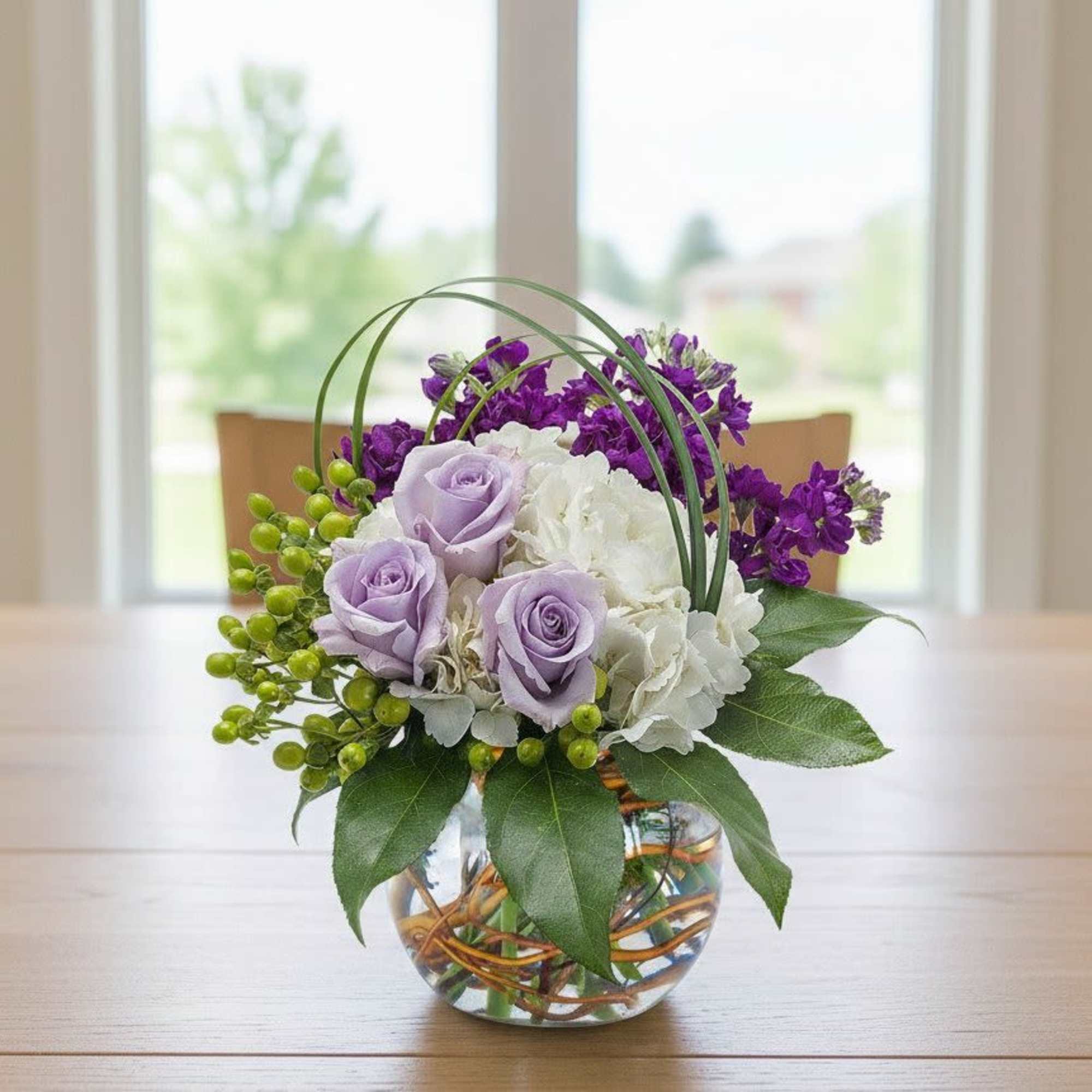 Round glass vase arrangement with lavender roses, white hydrangea, and purple flowers with green berries