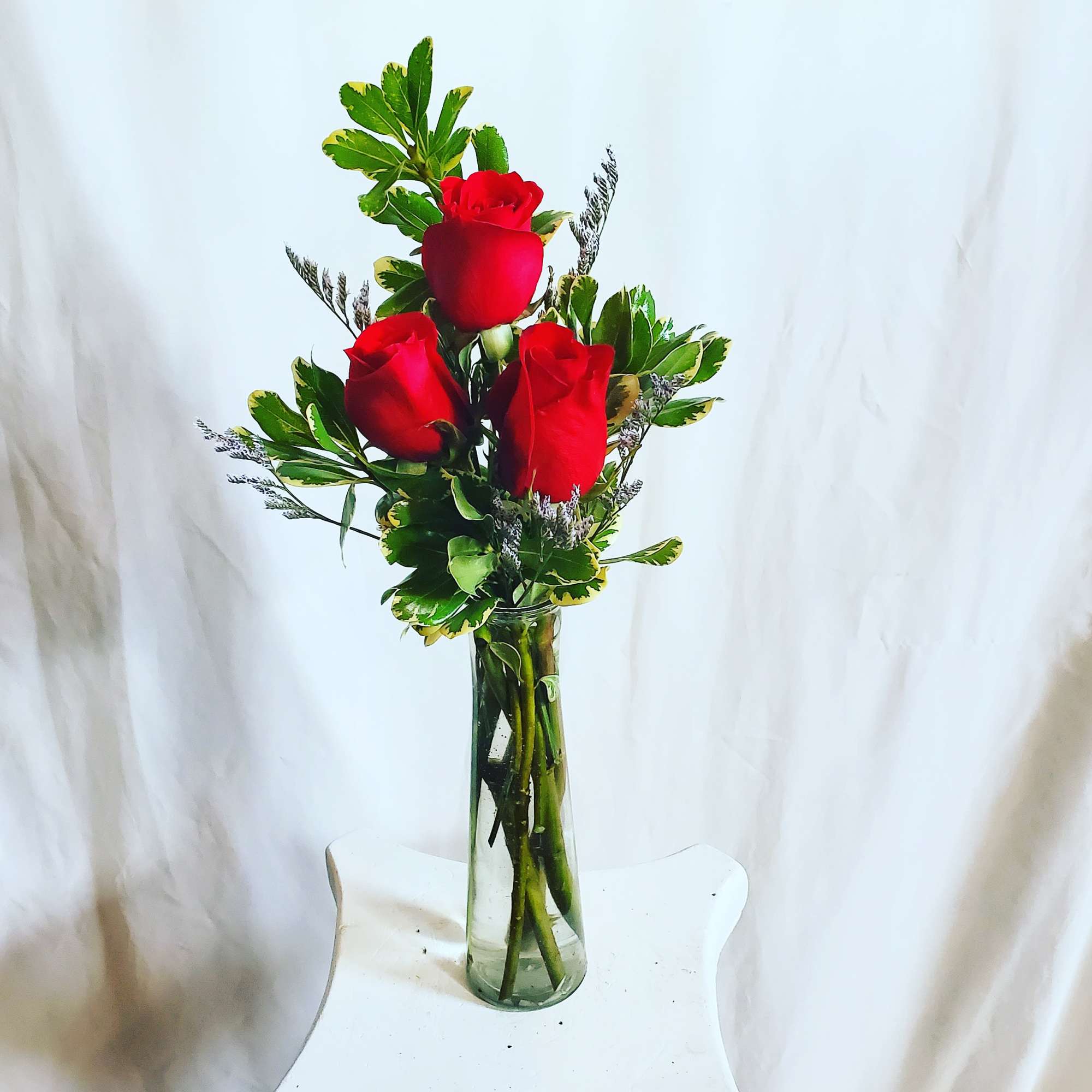 Three red roses in a clear glass vase with variegated greenery