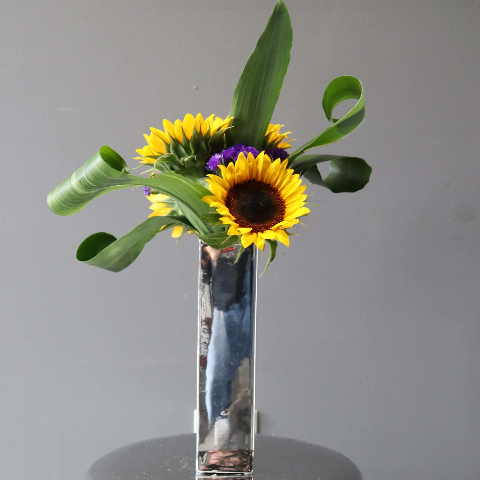 Sunflowers arranged in a tall mirrored vase with curled green leaves, photo 2 of 2