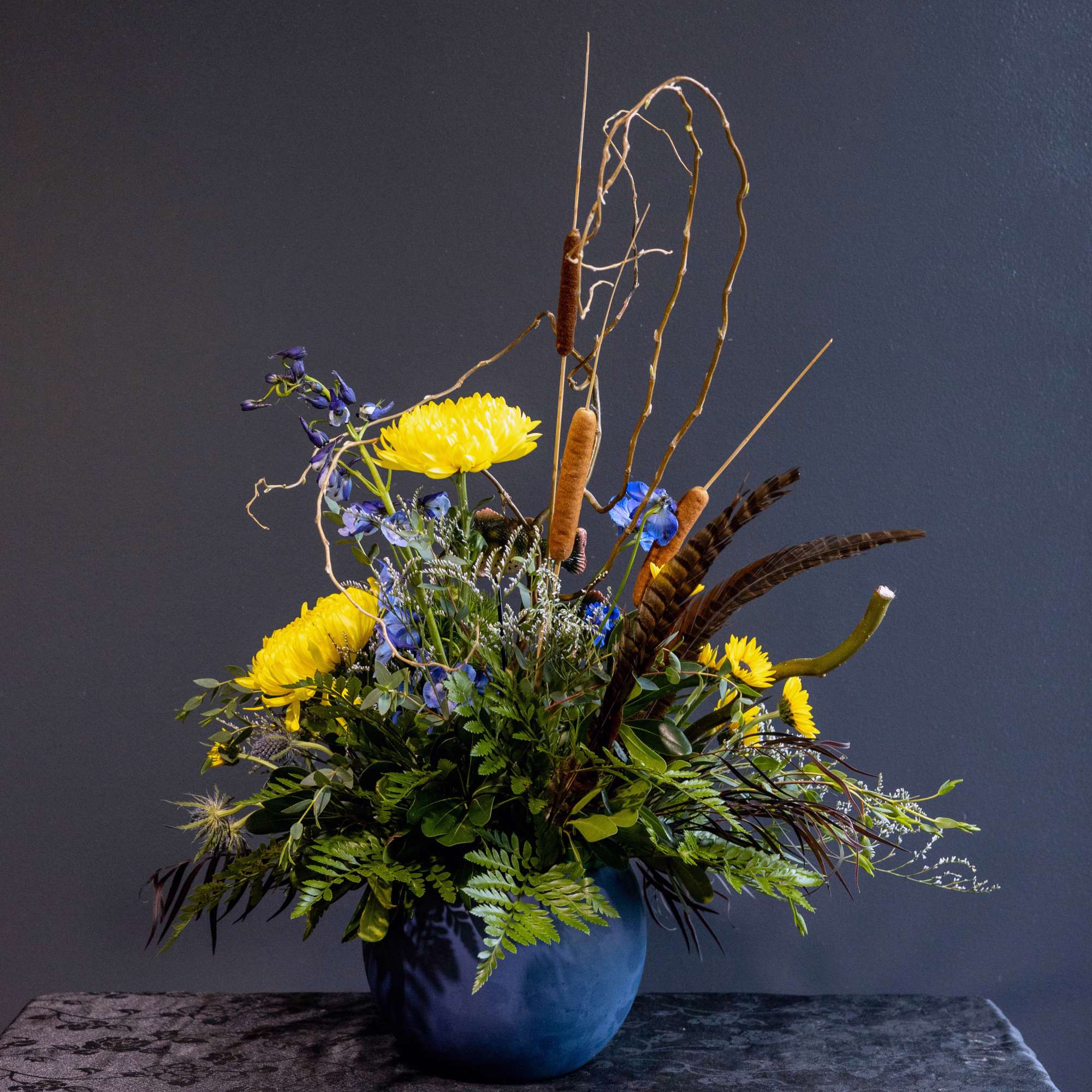 Yellow and blue floral arrangement in a blue vase with cattails and branches