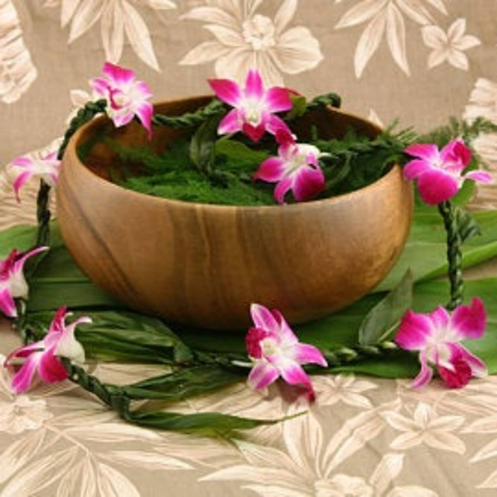 Pink orchid lei draped around a wooden bowl on a floral tablecloth