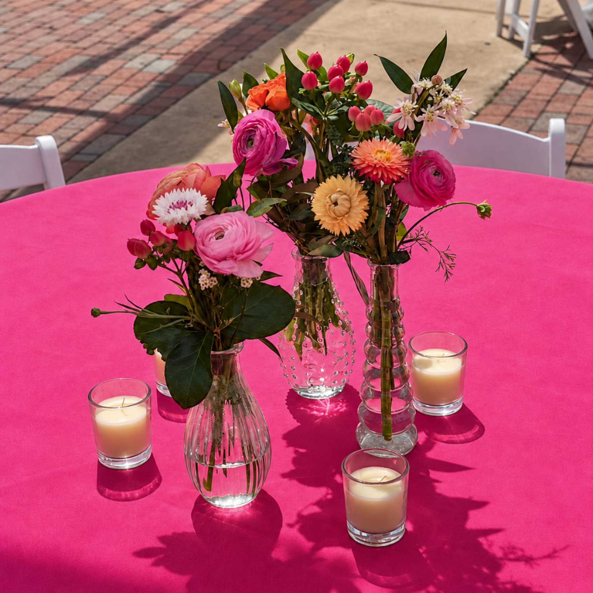 Three small floral arrangements in glass vases on a bright pink table with candles.