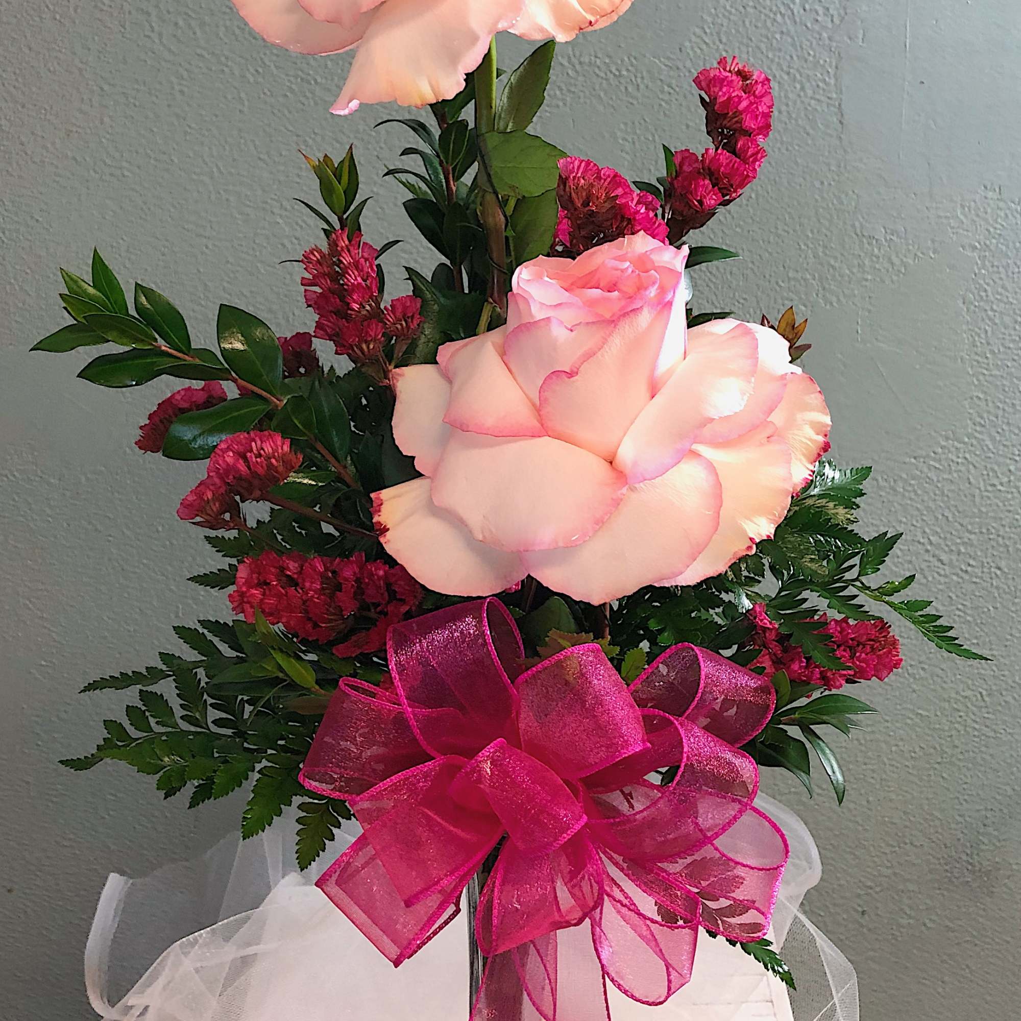 Tall bud vase with three pale pink roses, dark pink filler flowers, and a large hot pink ribbon bow.