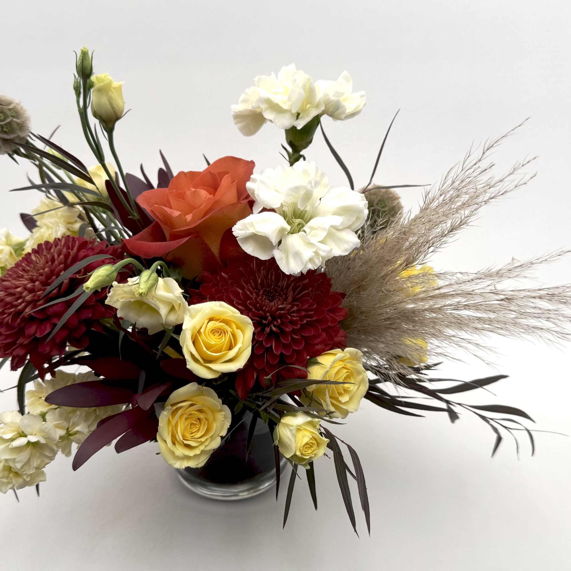 Low arrangement of red mums, yellow roses, white carnations, and pampas plumes in a clear glass vase.