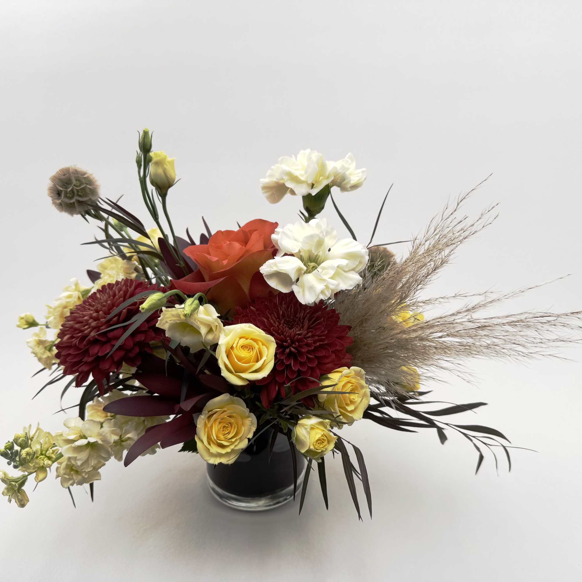 Low arrangement of red mums, yellow roses, white blooms and pampas grass in a round glass vase