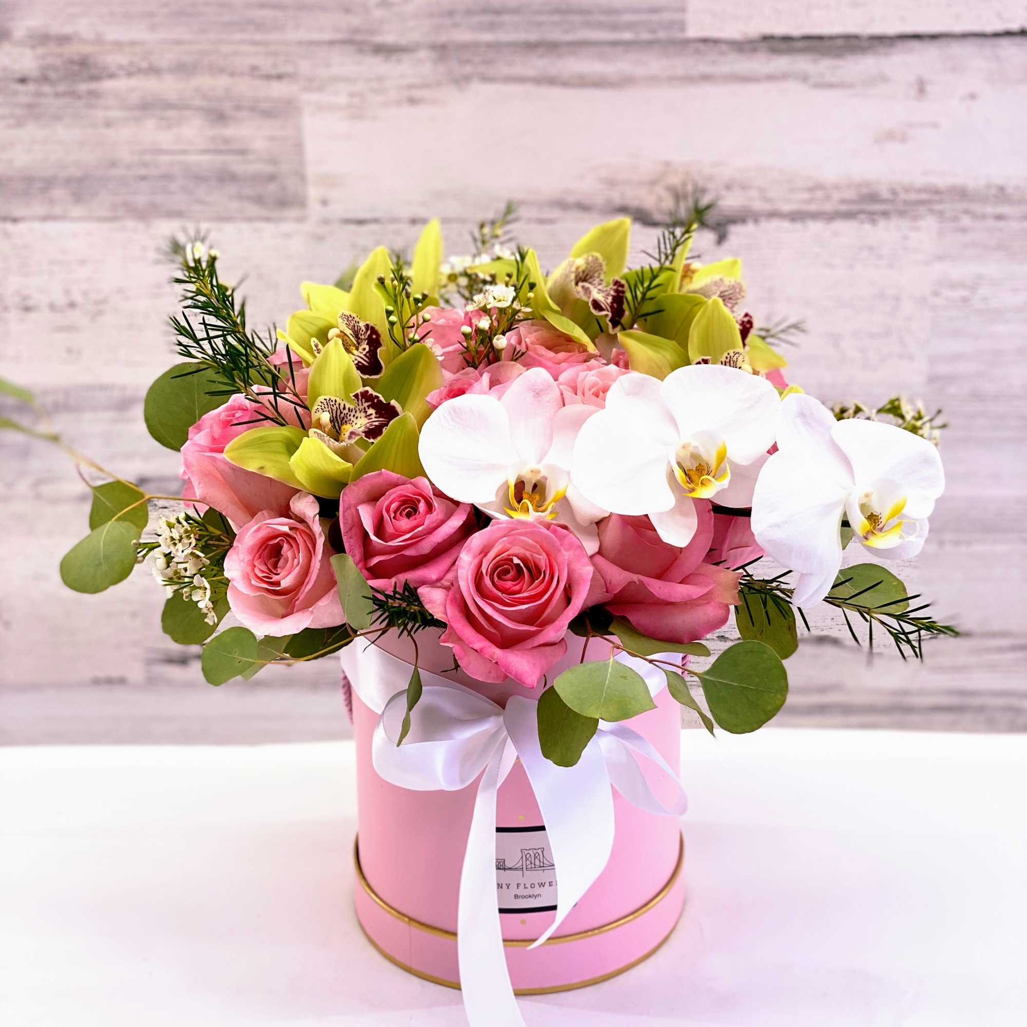 Pink roses and white orchids arranged in a pink hat box with a ribbon
