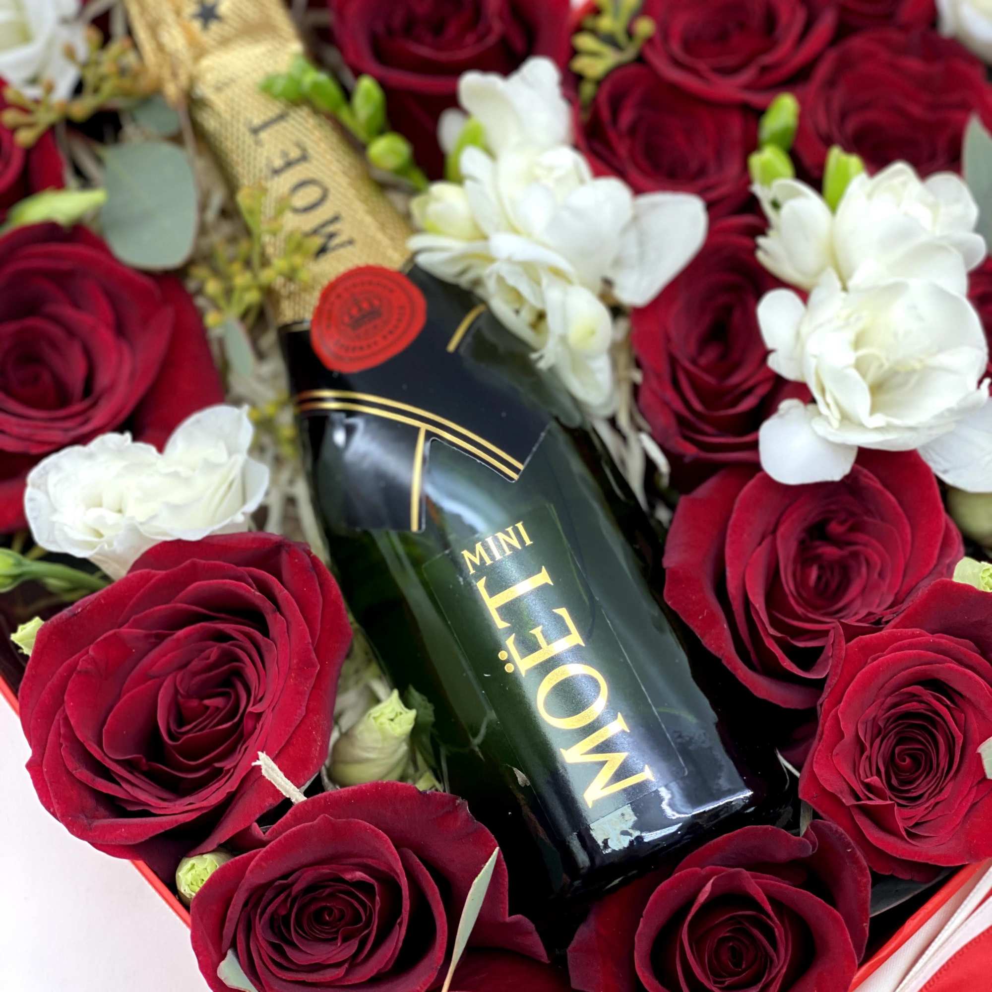 Red roses and white flowers arranged around a mini Moët bottle in a gift box
