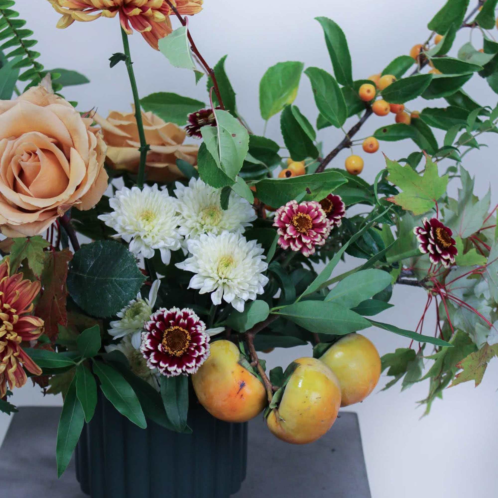 Mixed floral arrangement with peach roses, chrysanthemums, and fruit in a dark vase