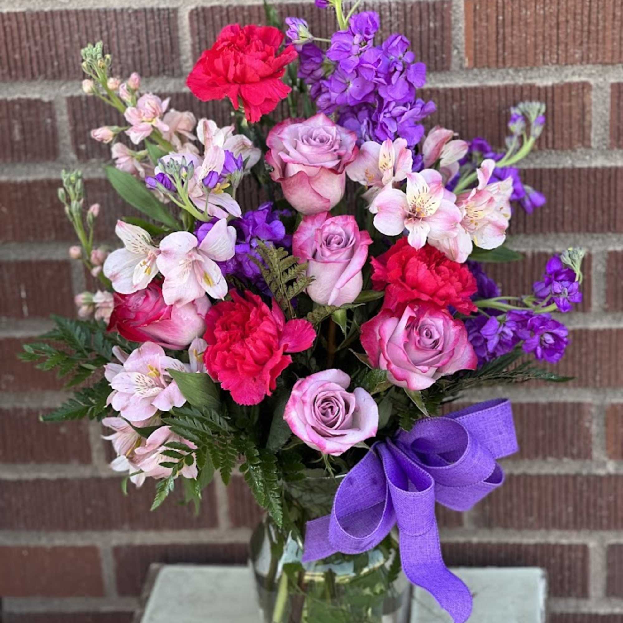 Bouquet of pink roses, carnations, and purple flowers in a glass vase with a ribbon