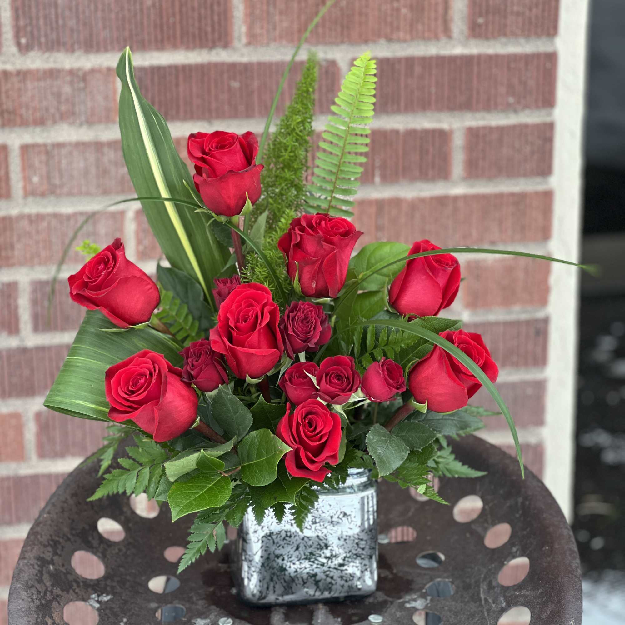 Red roses arranged in a square silver vase with mixed greenery