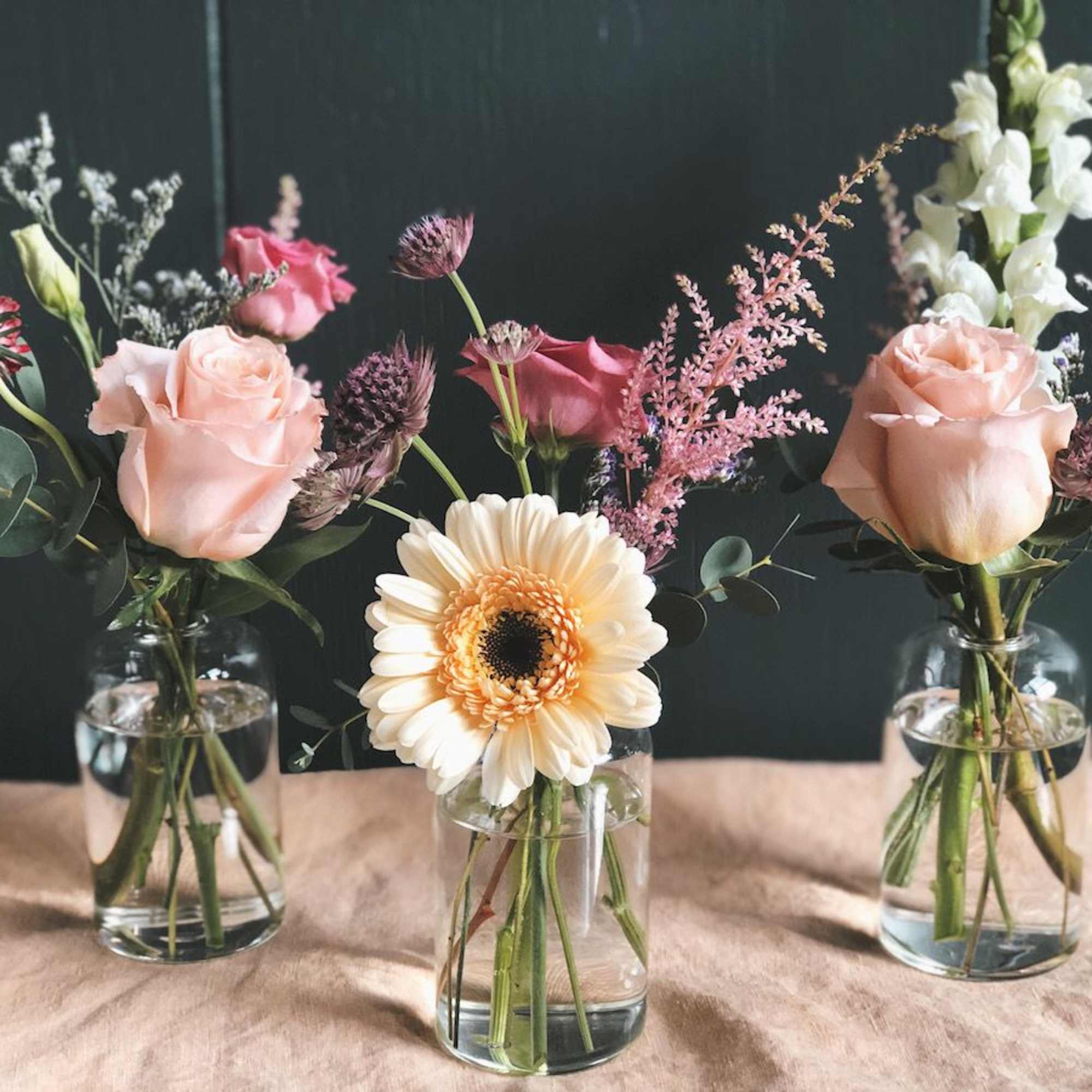Three small floral arrangements in glass jars with roses and a gerbera daisy