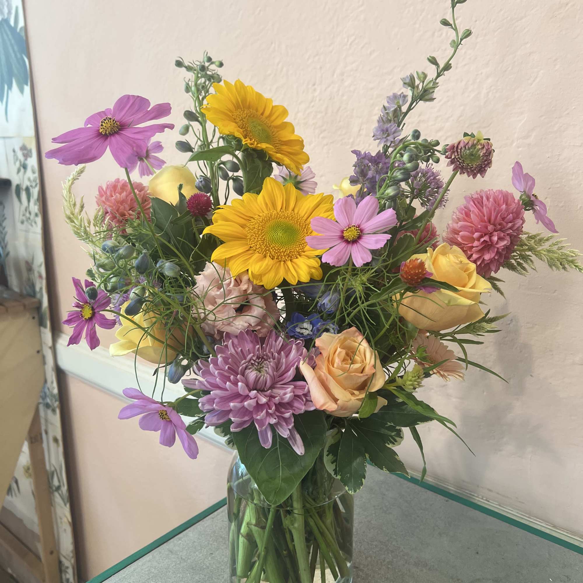 Mixed bouquet of yellow sunflowers, pink flowers, and peach roses in a glass vase
