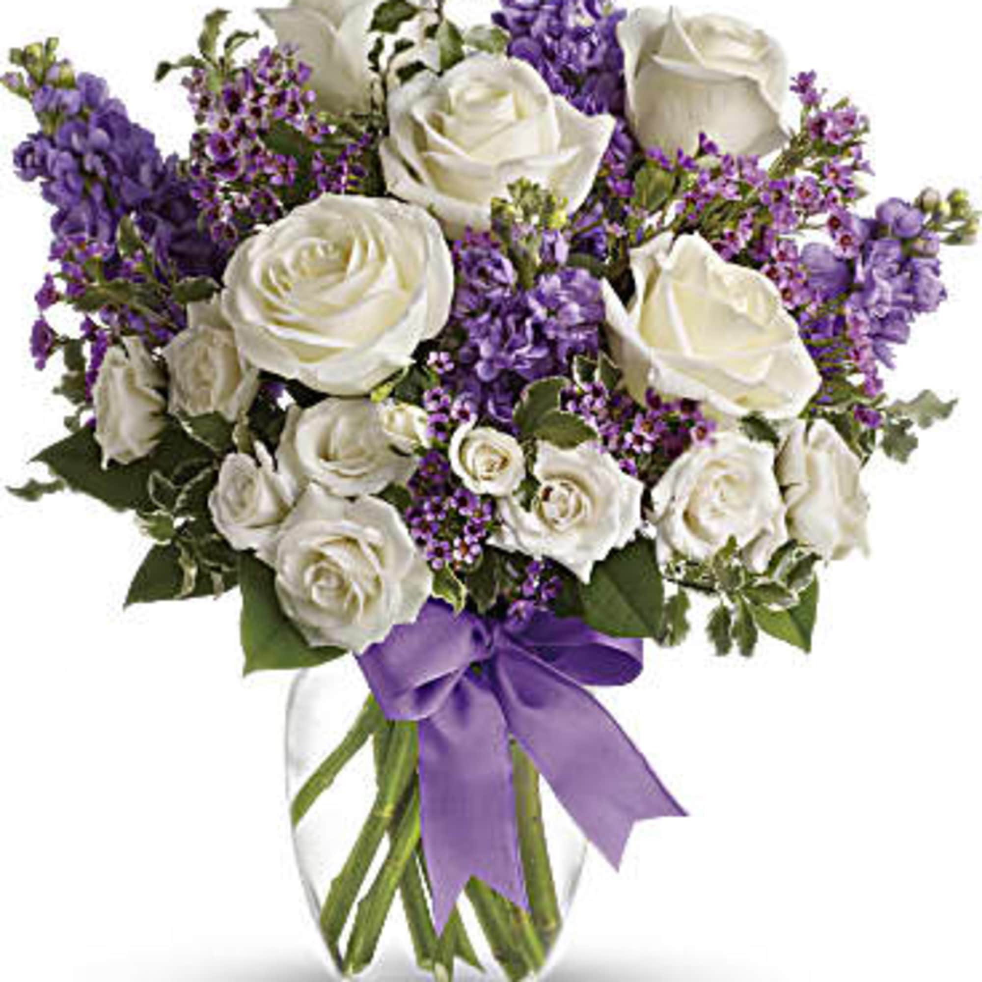 White roses and purple flowers arranged in a glass vase with a purple ribbon