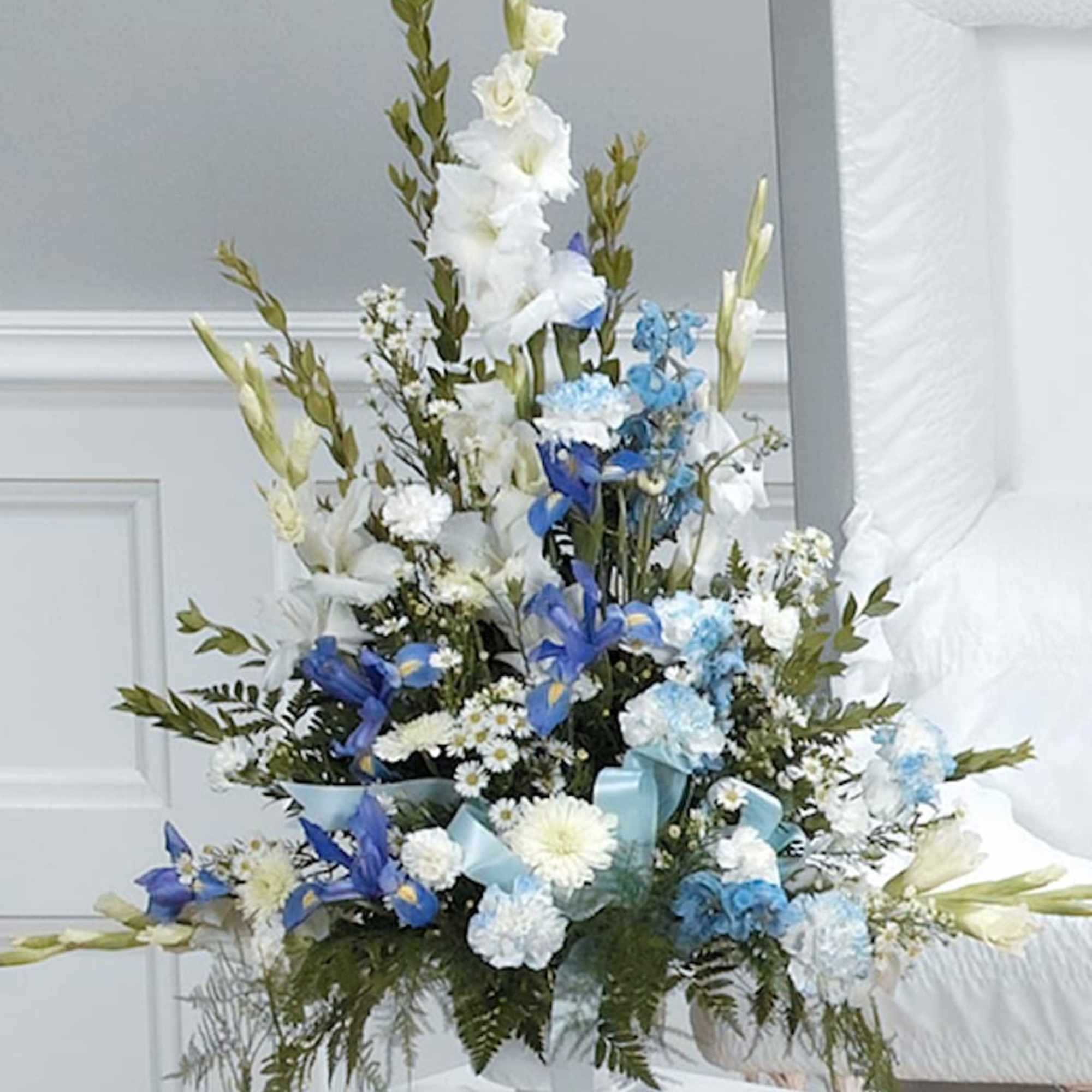 Tall blue and white sympathy flower arrangement in a white pedestal vase beside a casket