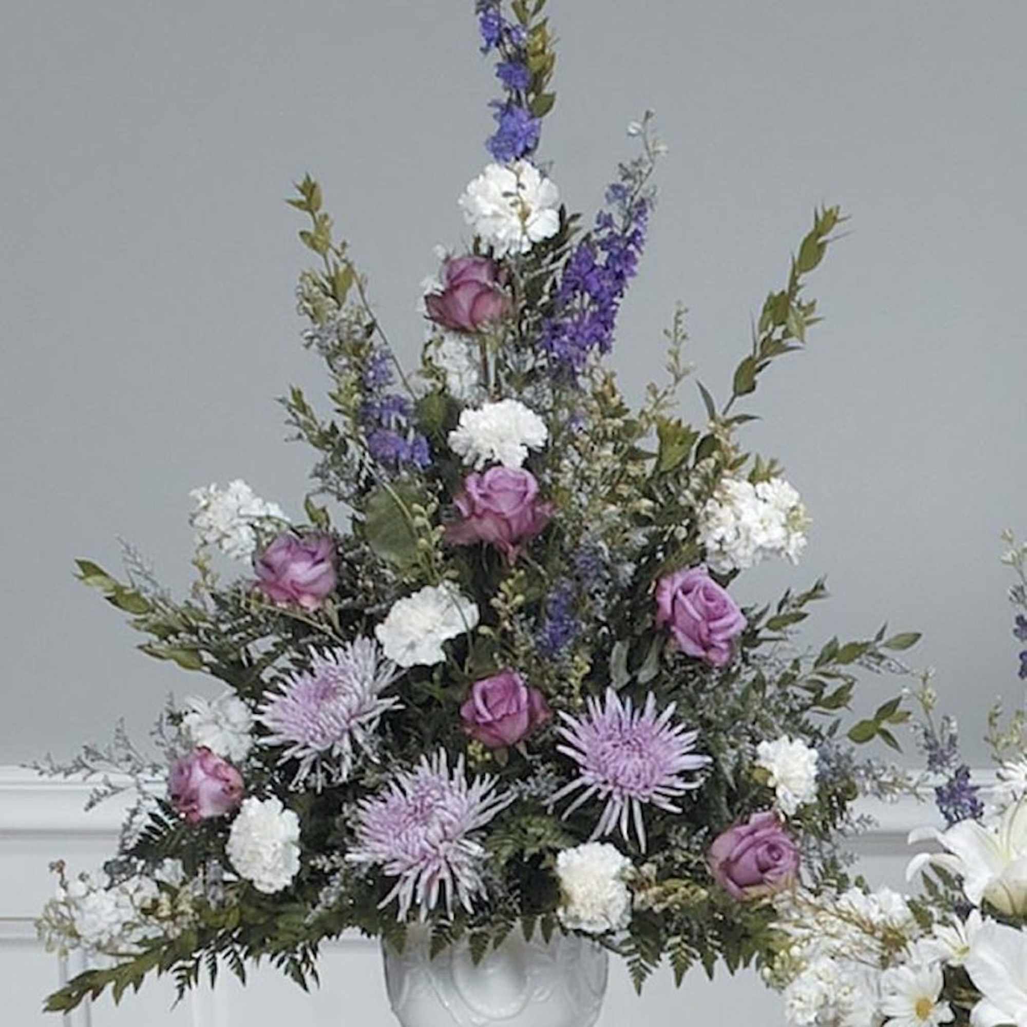 Tall lavender and white funeral-style flower arrangement in a white pedestal urn