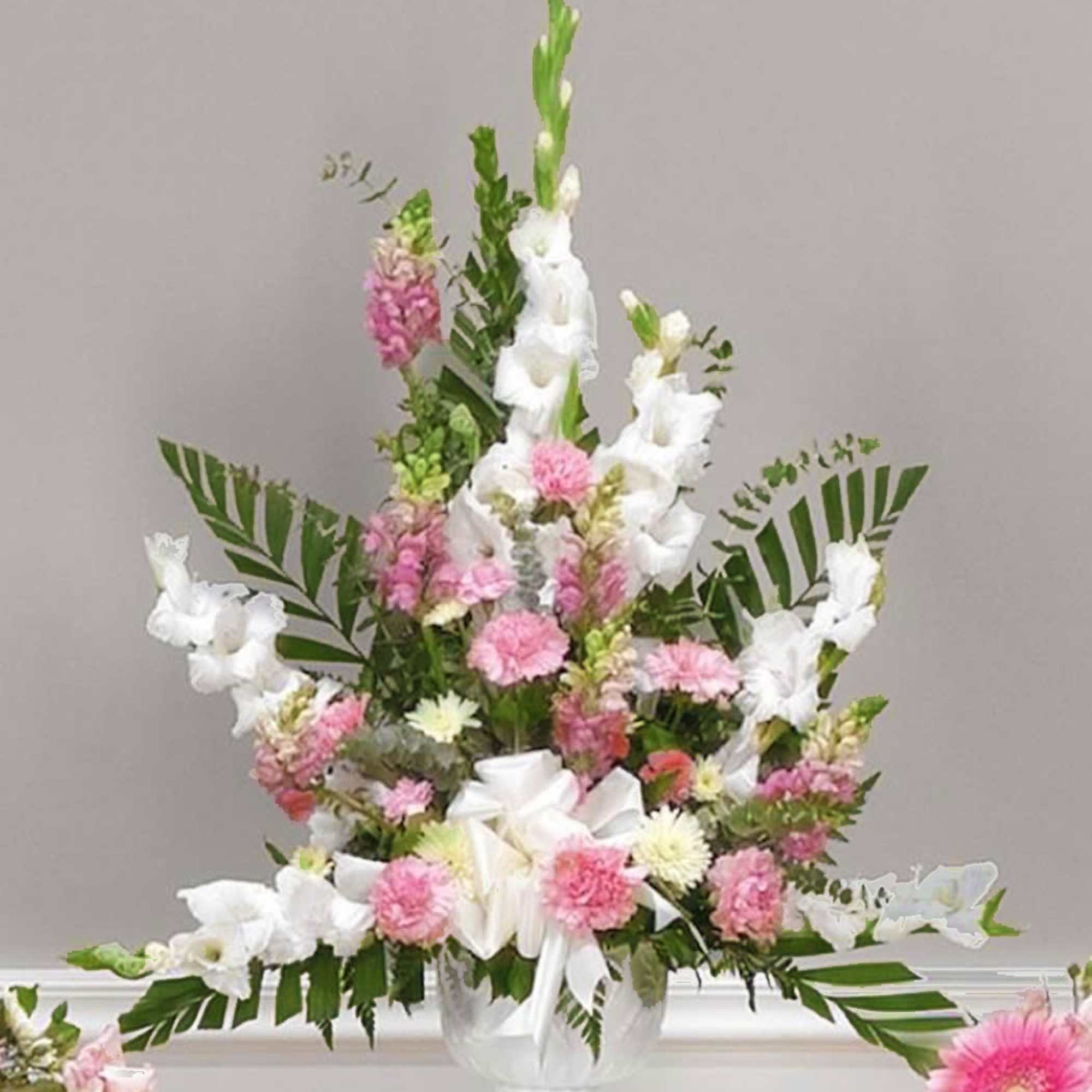 Tall pink and white arrangement with carnations and gladiolus in a white urn with a bow