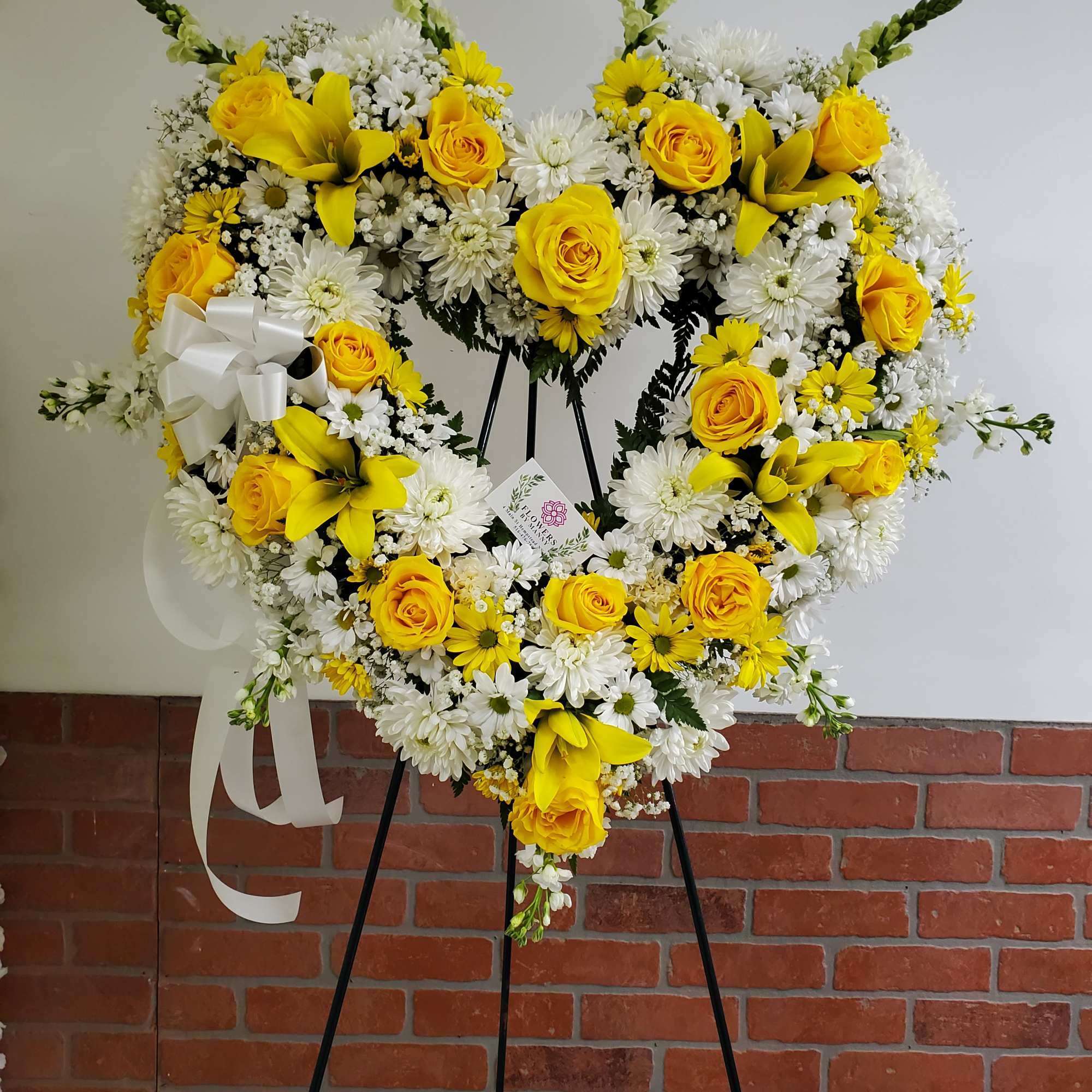 Heart-shaped standing wreath of yellow roses, white daisies and lilies with a white bow on an easel.
