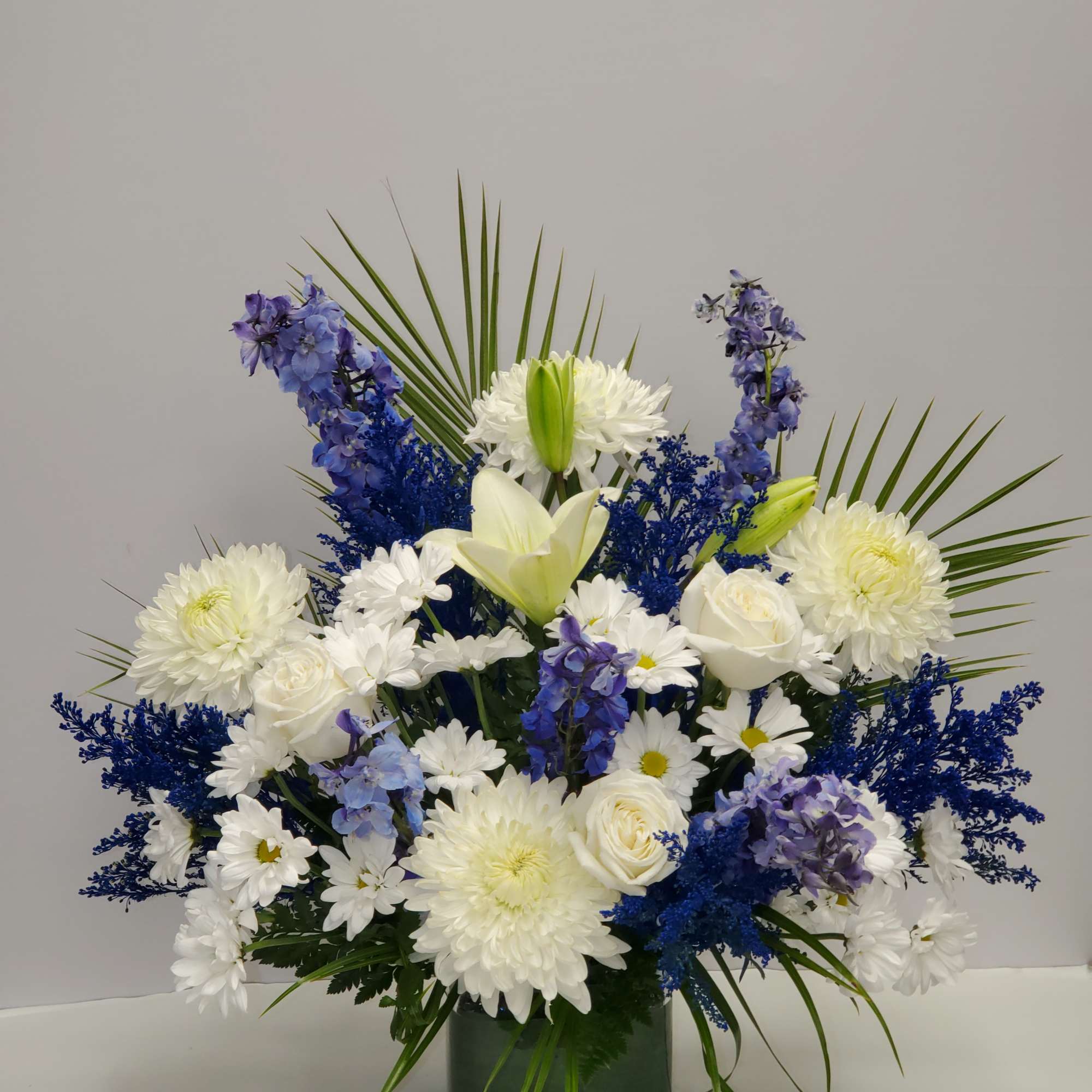 Tall arrangement of white lilies, roses, mums, and blue flowers in a green glass vase