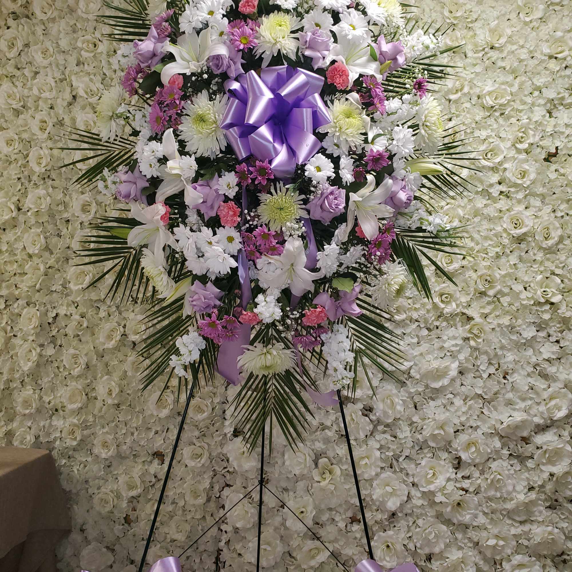 Tall standing spray of white, lavender, and pink flowers with a large lavender bow on an easel.