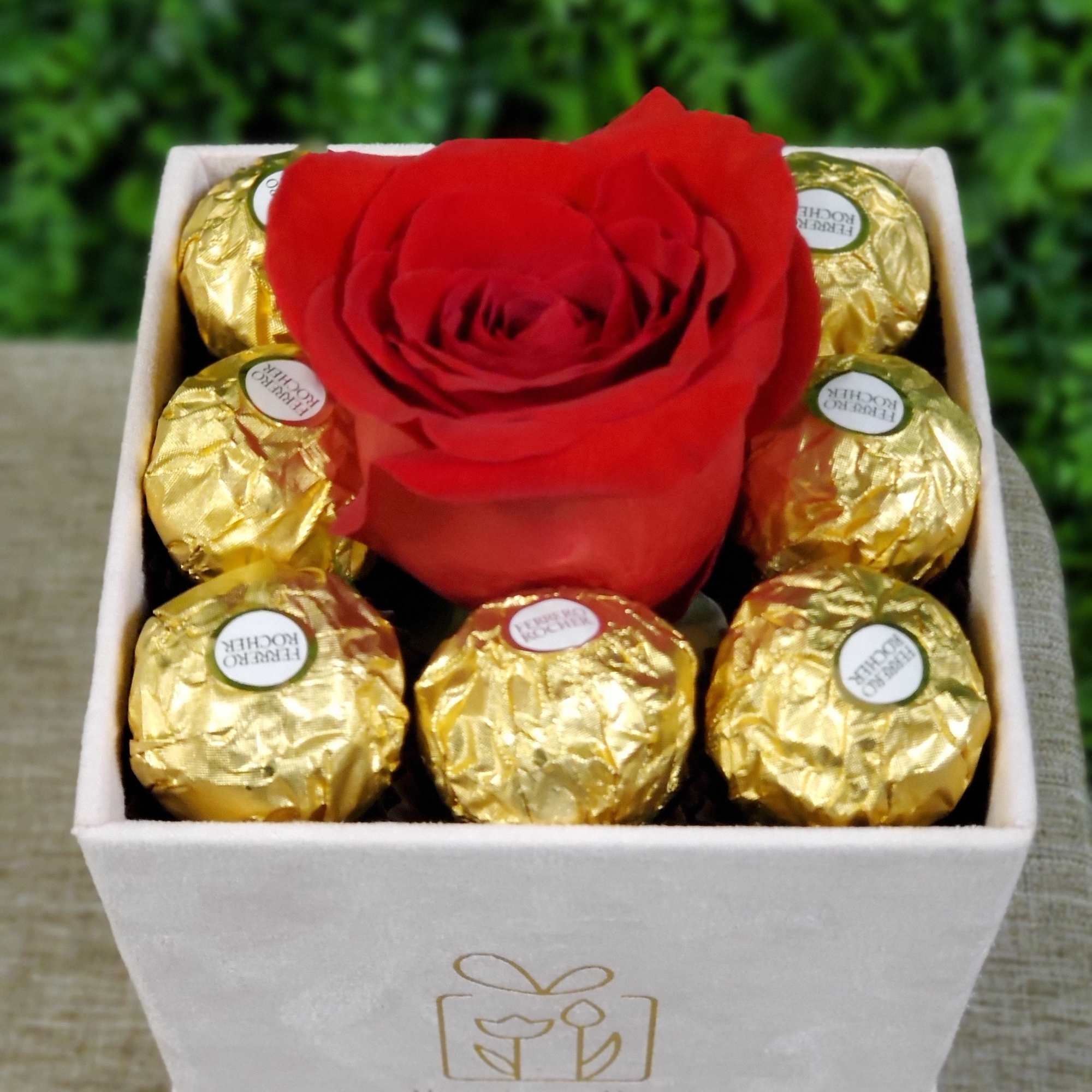 Square box with a single red rose surrounded by gold-wrapped chocolates