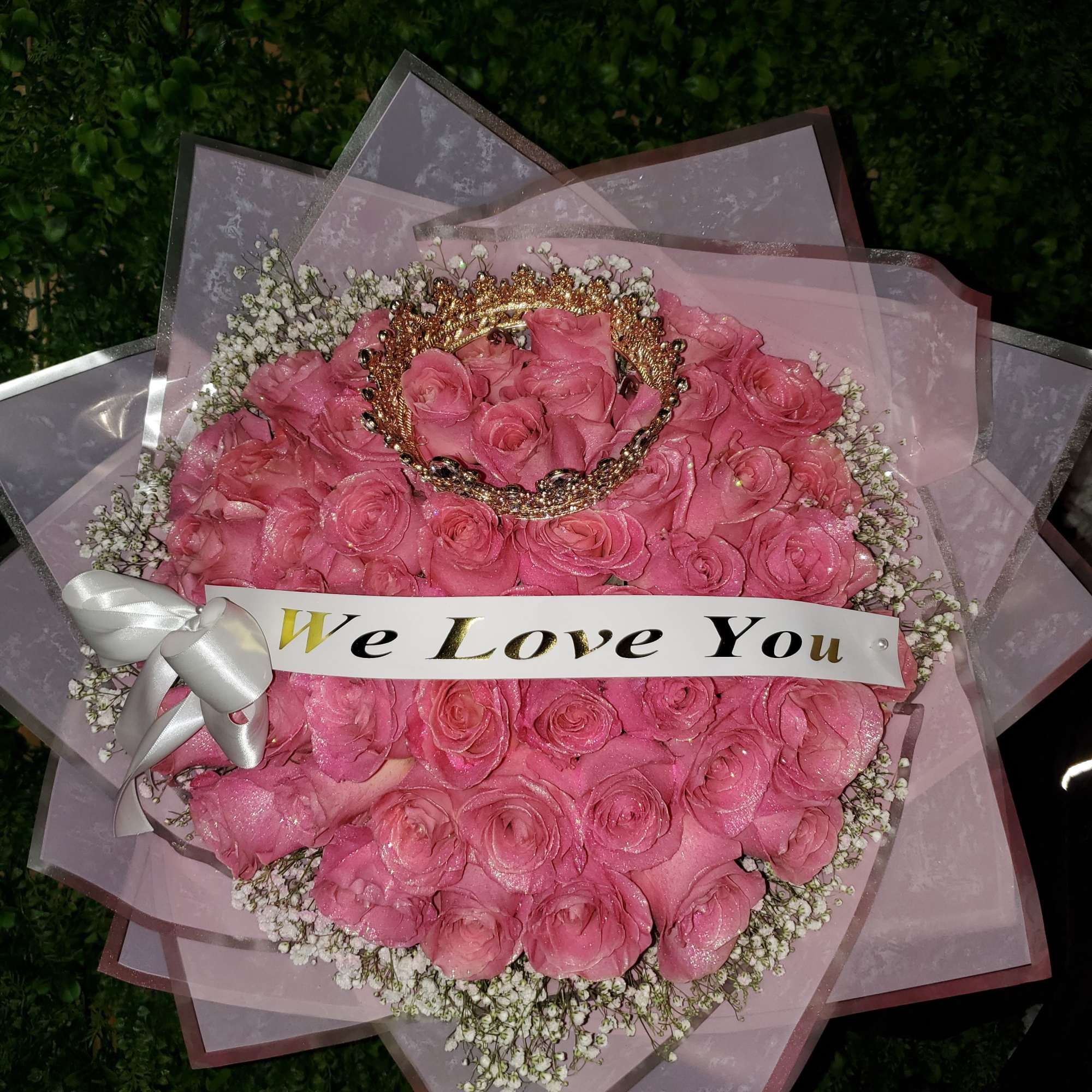 Round bouquet of pink roses with baby’s breath, crown accent, and a ‘We Love You’ banner.