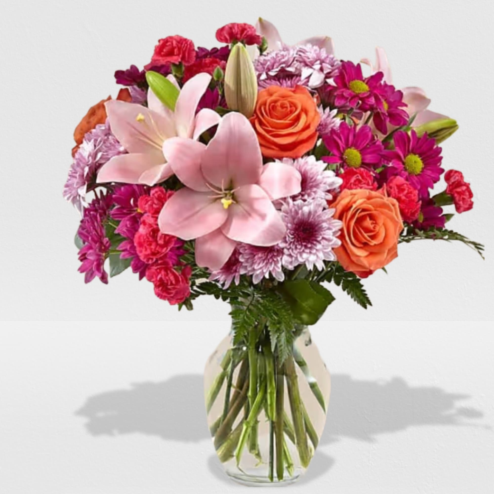Mixed bouquet of pink lilies, orange roses, and purple mums in a clear glass vase