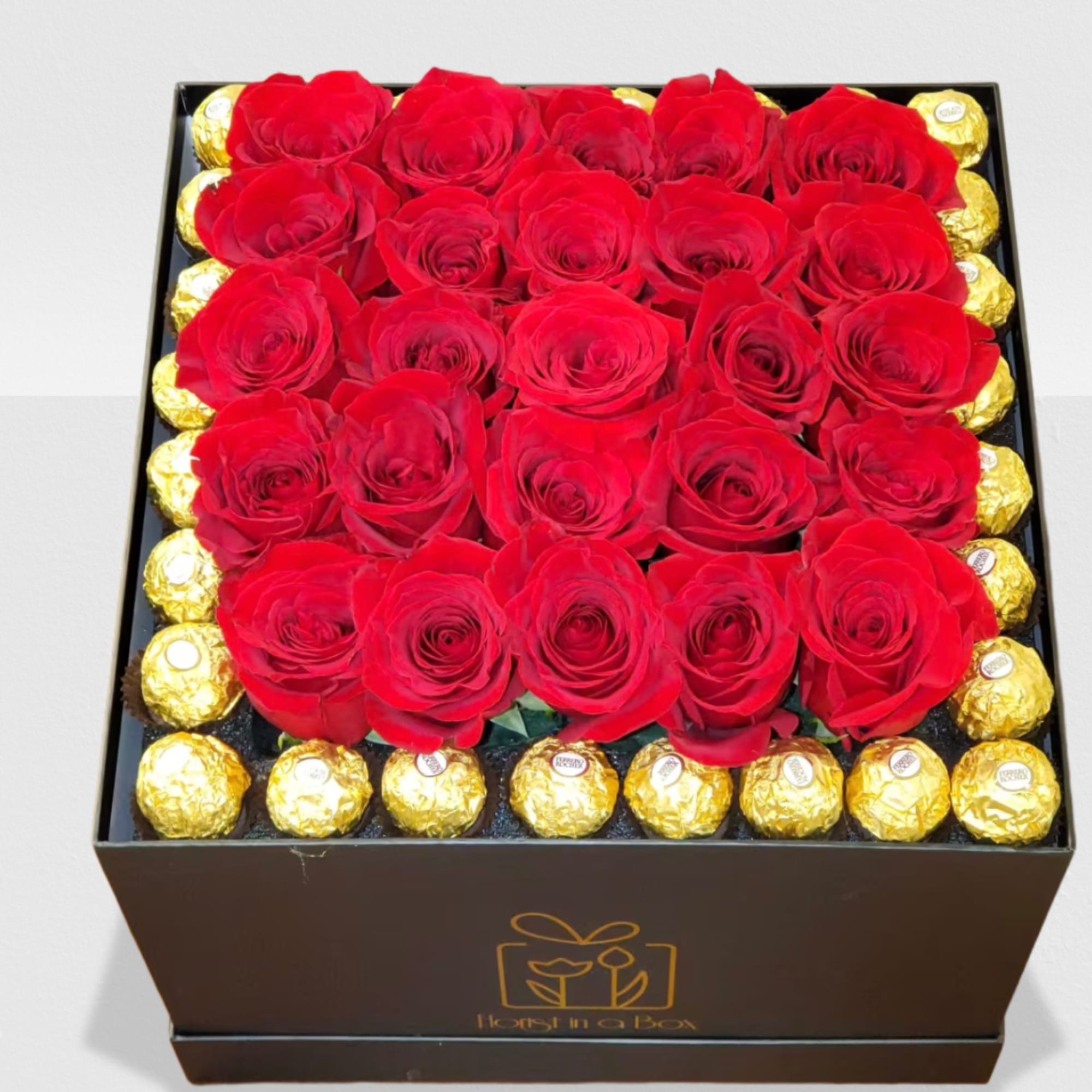 Square black box filled with red roses surrounded by gold-wrapped chocolates